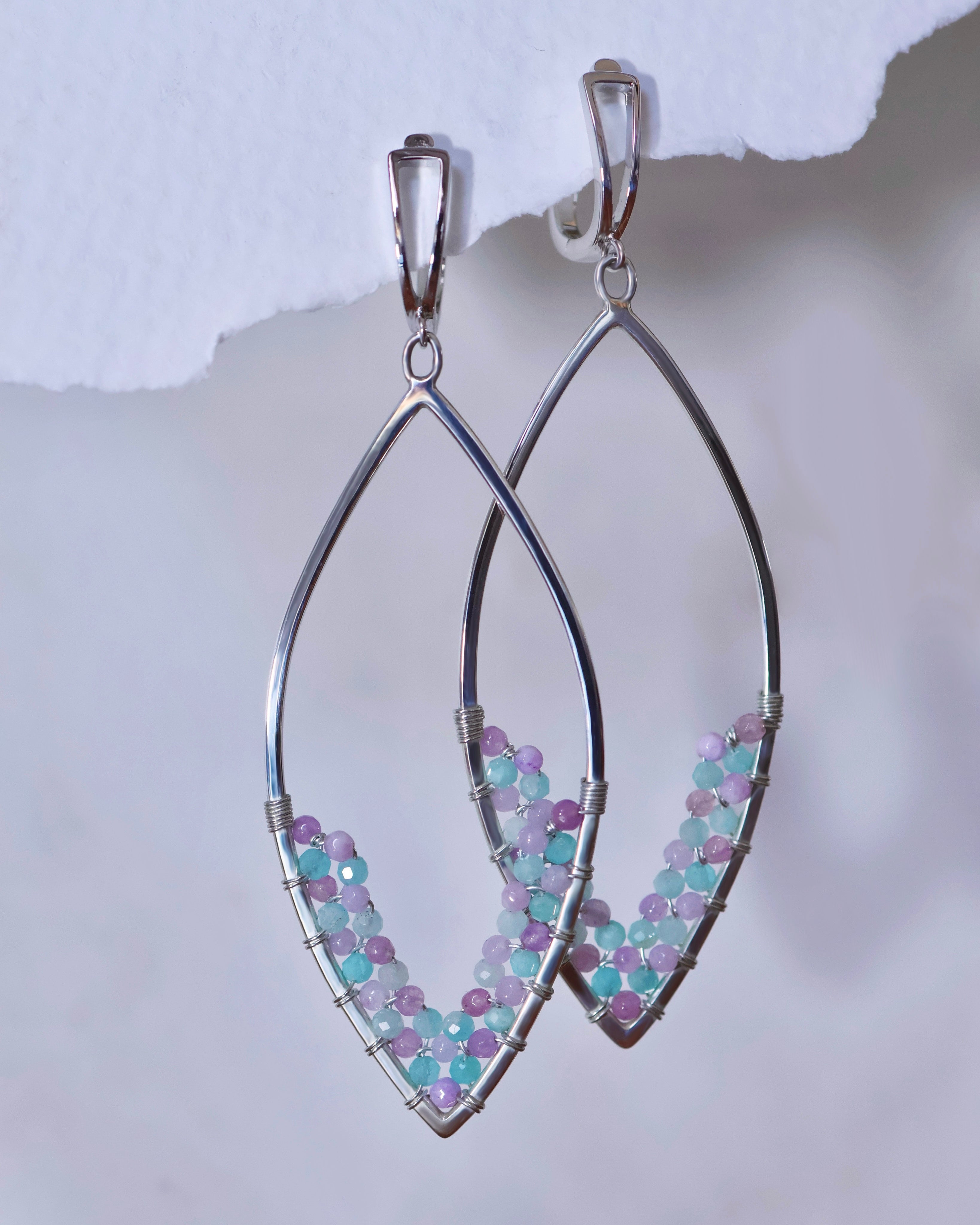 The Amethyst + Amazonite Earrings by Lilac Sky Studio are sterling silver drop earrings with an open marquise shape, featuring woven amethyst and amazonite beads along the lower half.