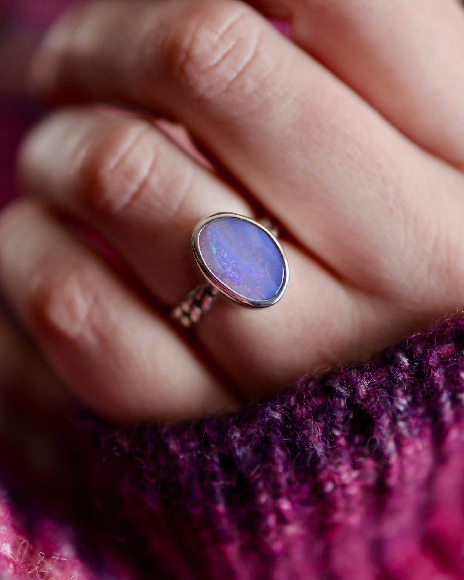 A hand wears the Lilac Sky Studio Boulder Opal Ring, featuring a sterling silver band and an oval purple gemstone, resting on purple knit fabric.