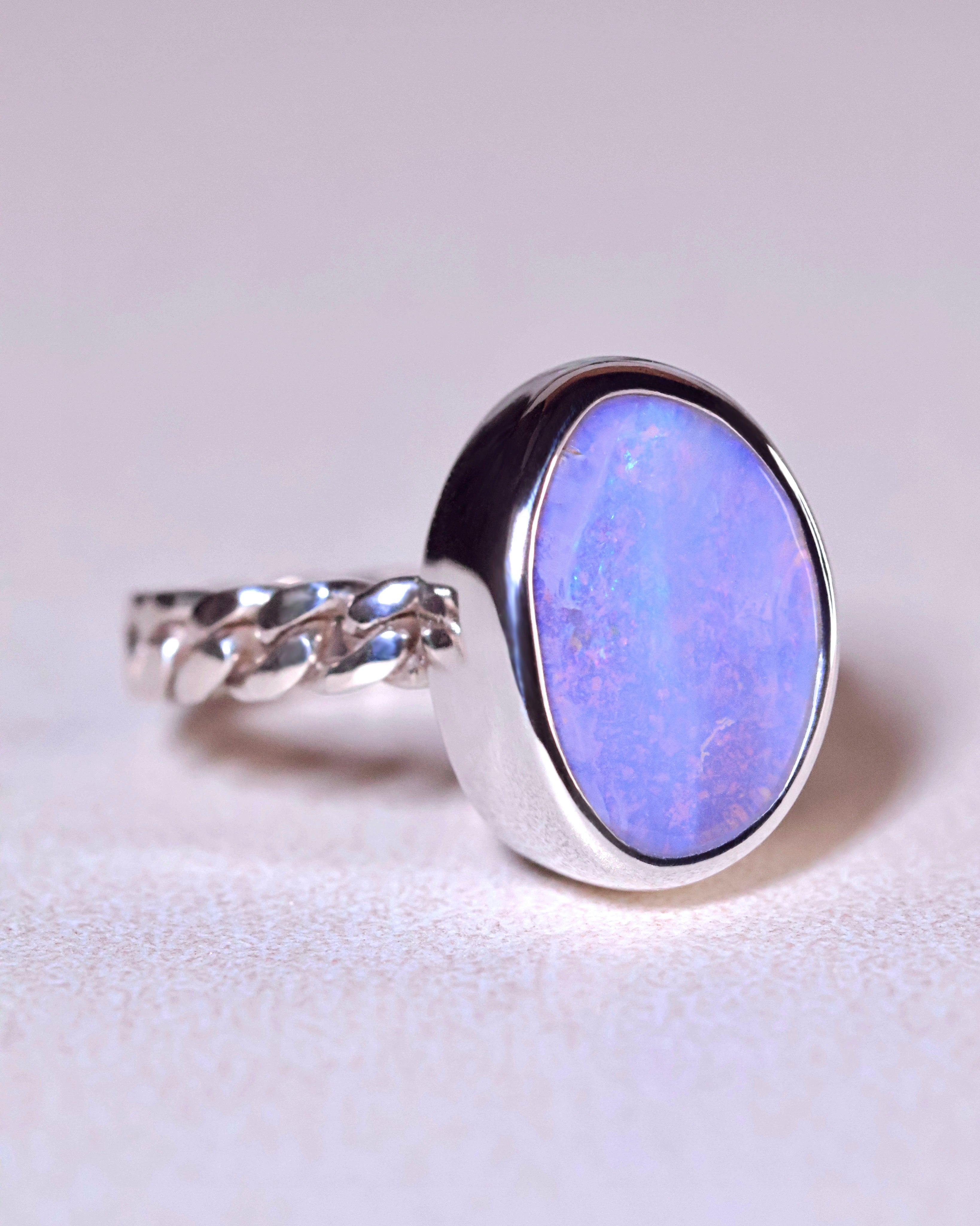 The Boulder Opal Ring by Lilac Sky Studio features an iridescent blue opal in a smooth bezel setting on a twisted sterling silver band, displayed on a neutral surface.