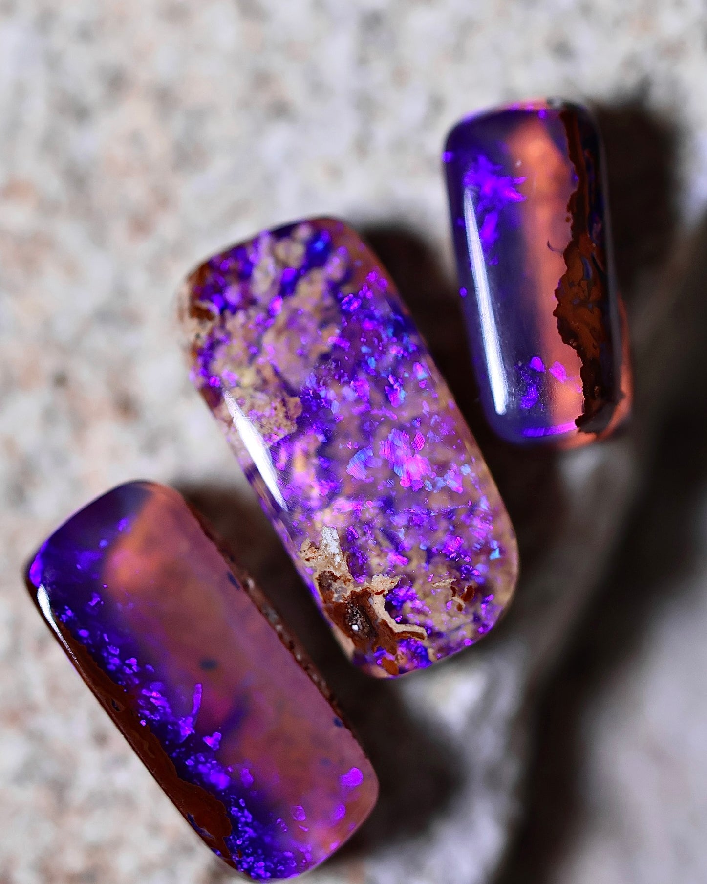 Three polished opal stones with purple and blue iridescent patterns are displayed on a textured stone surface, perfect for ethically sourced jewelry or to accent gold settings.