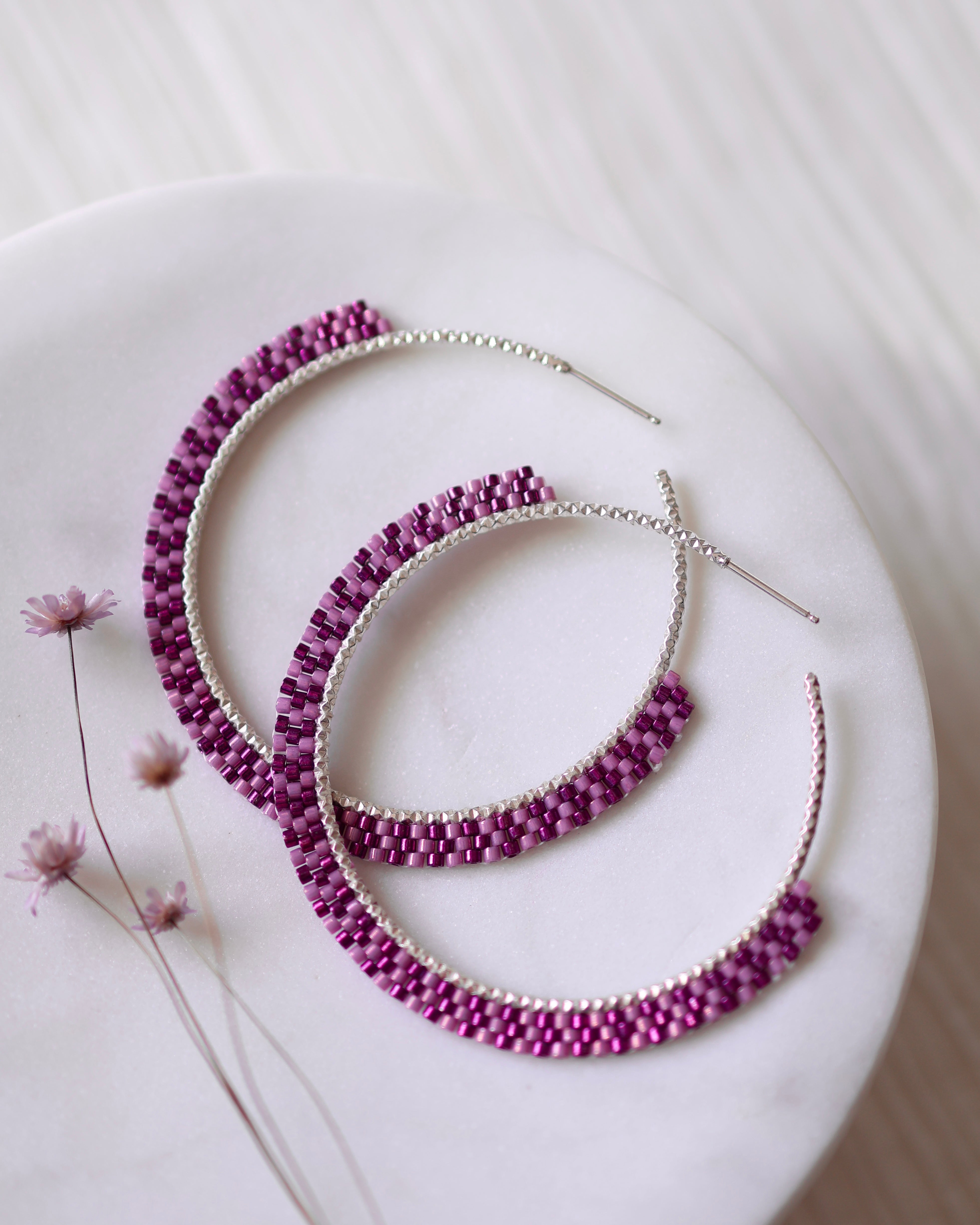 The Magenta Magic Hoops by Lilac Sky Studio—sterling silver handwoven hoops with magenta Japanese glass seed beads—are displayed on a round white surface, accompanied by delicate dried flowers.