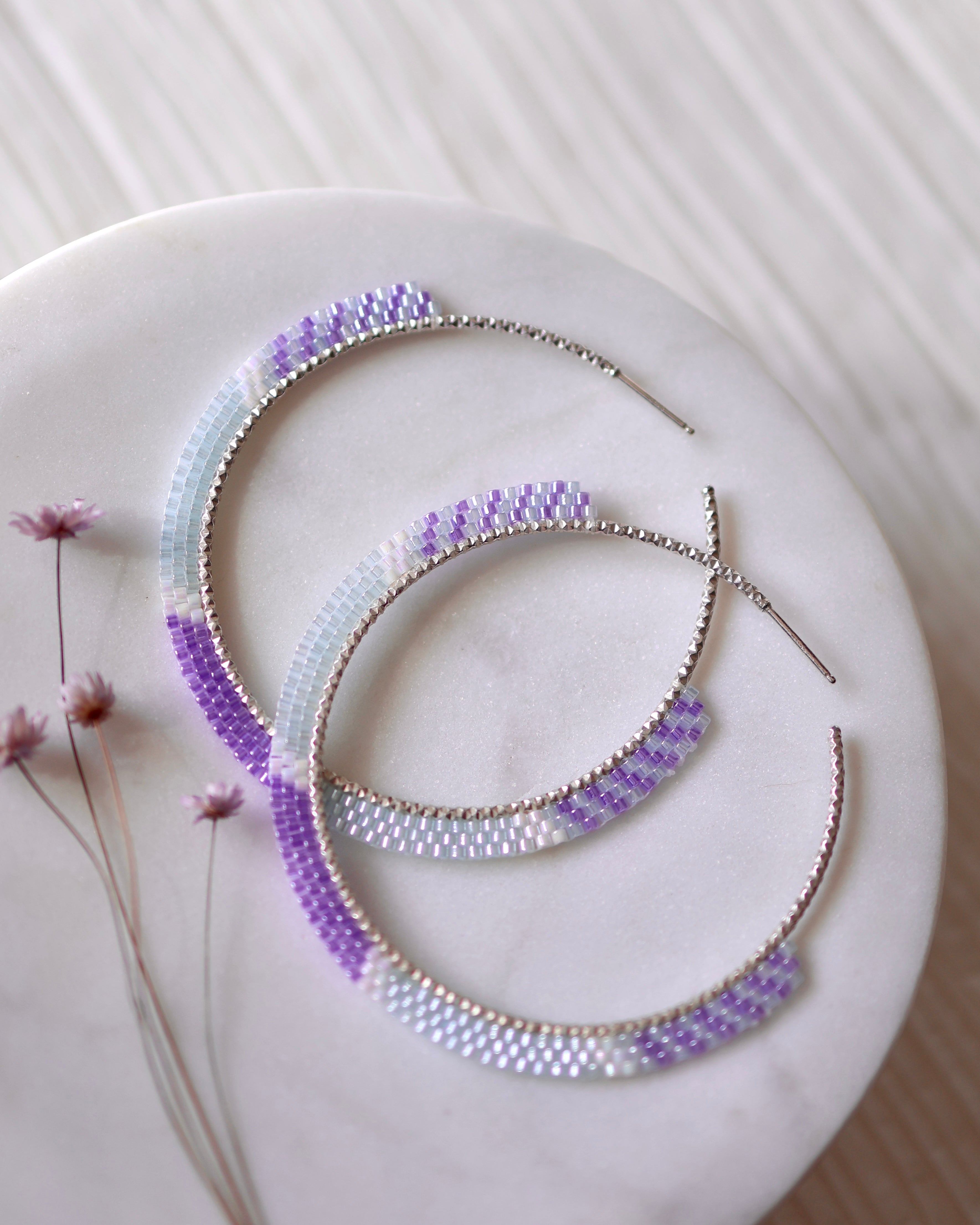 The Dreamweaver Hoops by Lilac Sky Studio are large statement earrings, handwoven with Japanese glass seed beads in a gradient of white, light blue, and lavender. Shown on a round white surface beside dried flowers.