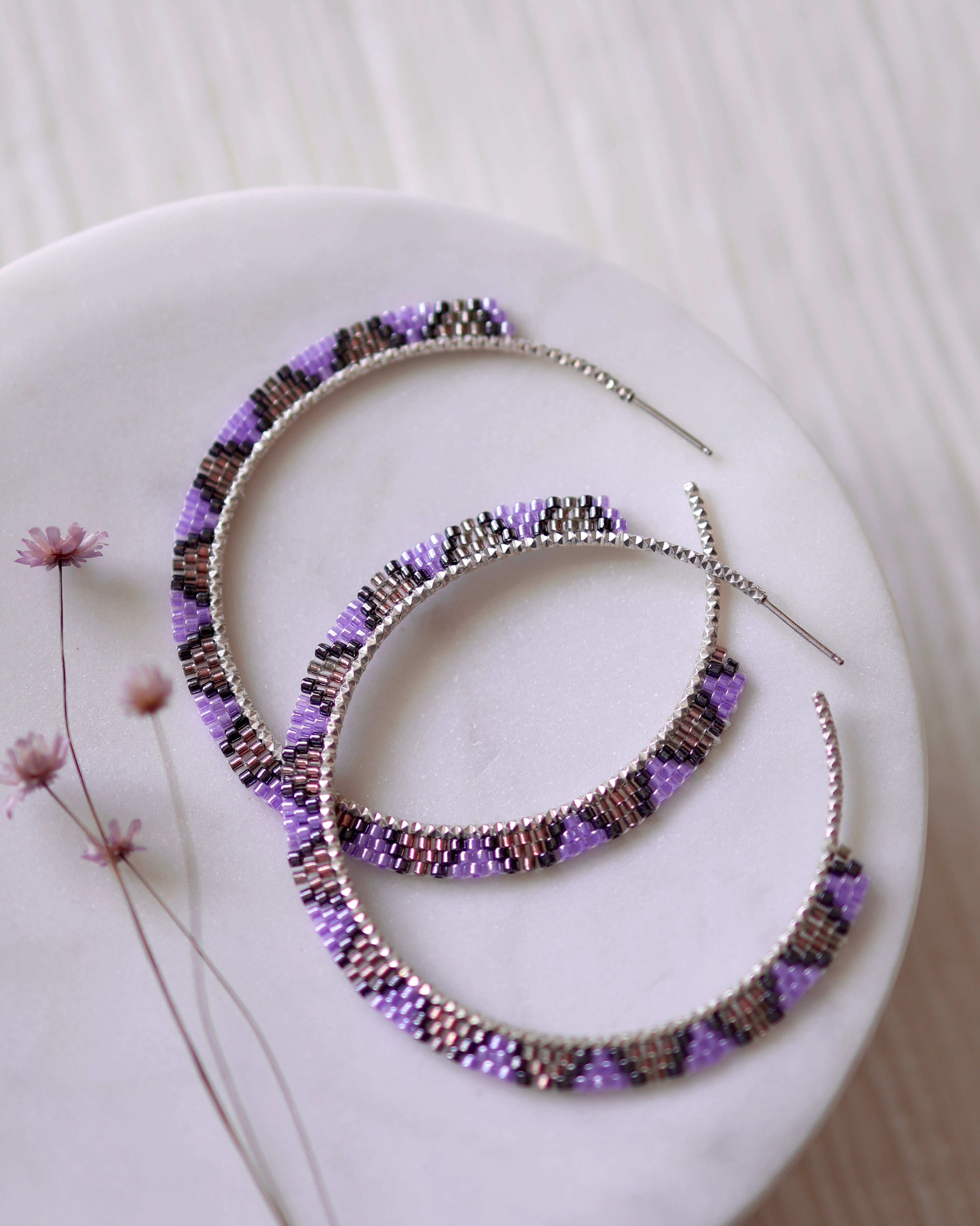 Purple Dusk Hoops by Lilac Sky Studio are large, handwoven sterling silver earrings with alternating purple, silver, and dark Japanese glass seed beads, beautifully showcased on white marble next to delicate dried flowers.