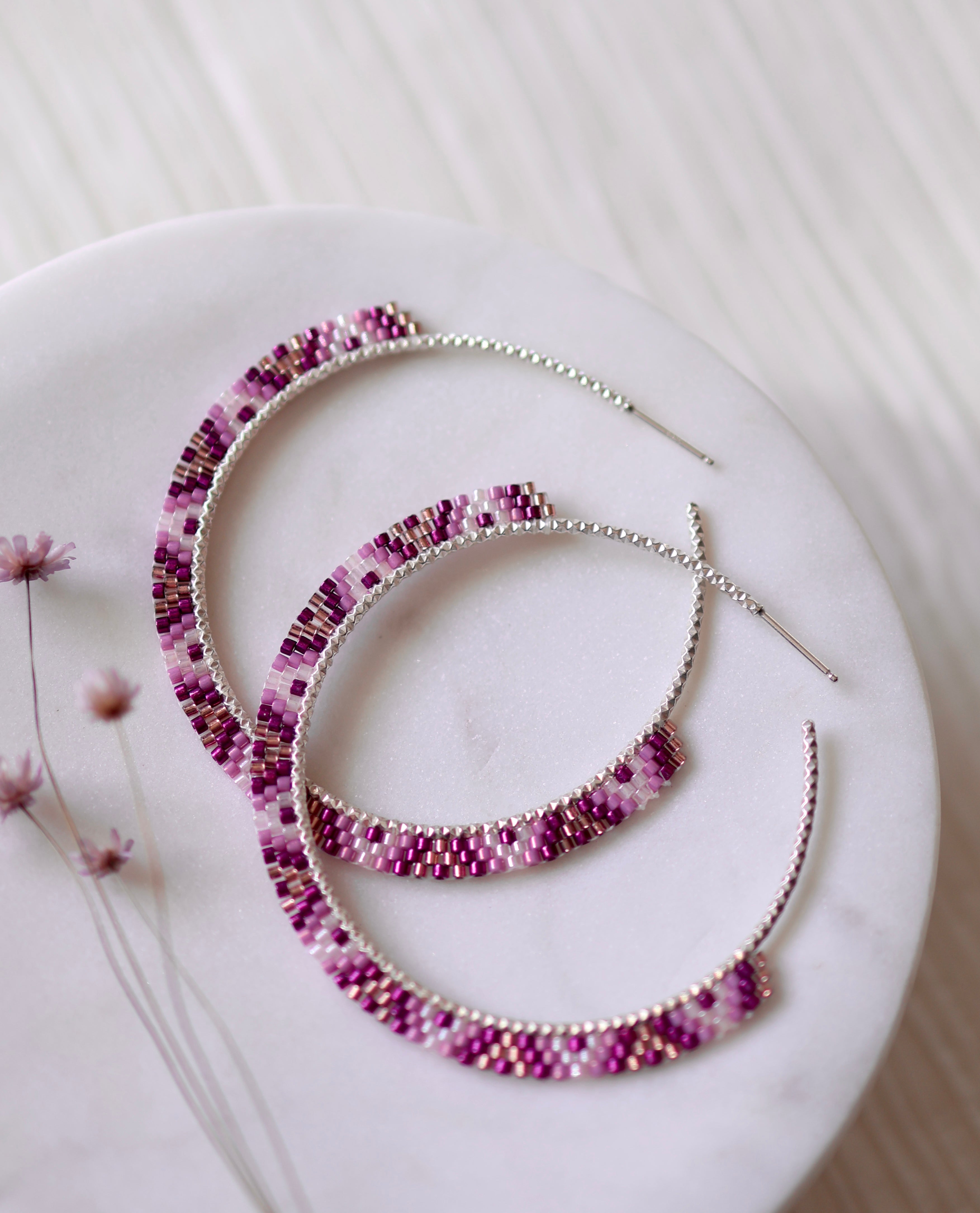 The Sunset Glow Hoops by Lilac Sky Studio are large curved handwoven earrings featuring a pink and purple beaded pattern with Japanese glass seed beads, displayed on a round white surface accented by small dried flowers.