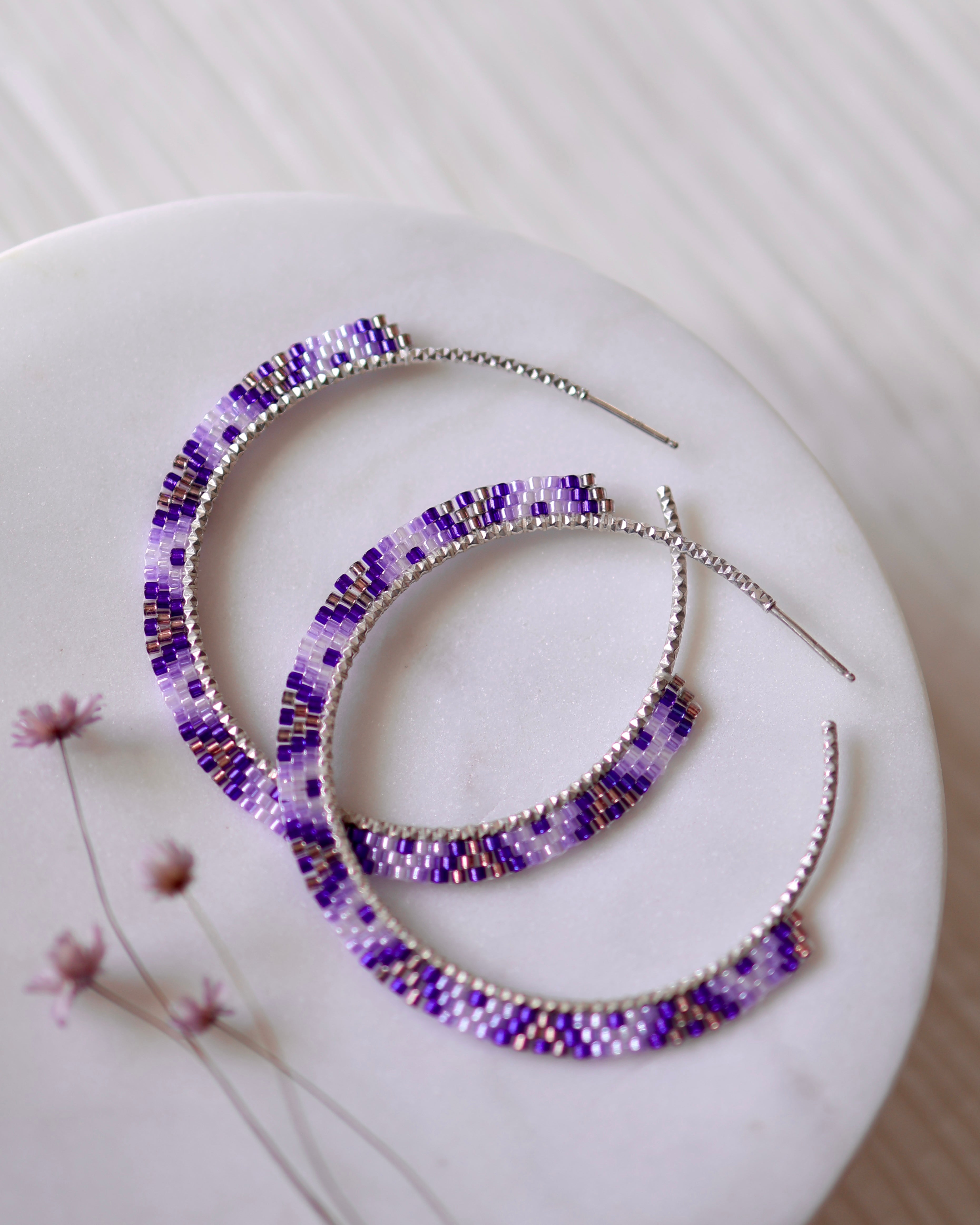 Violet Dreams Hoops by Lilac Sky Studio: sterling silver earrings adorned with purple, white, and gold Japanese glass seed beads, displayed on a round white surface beside tiny pink flowers.