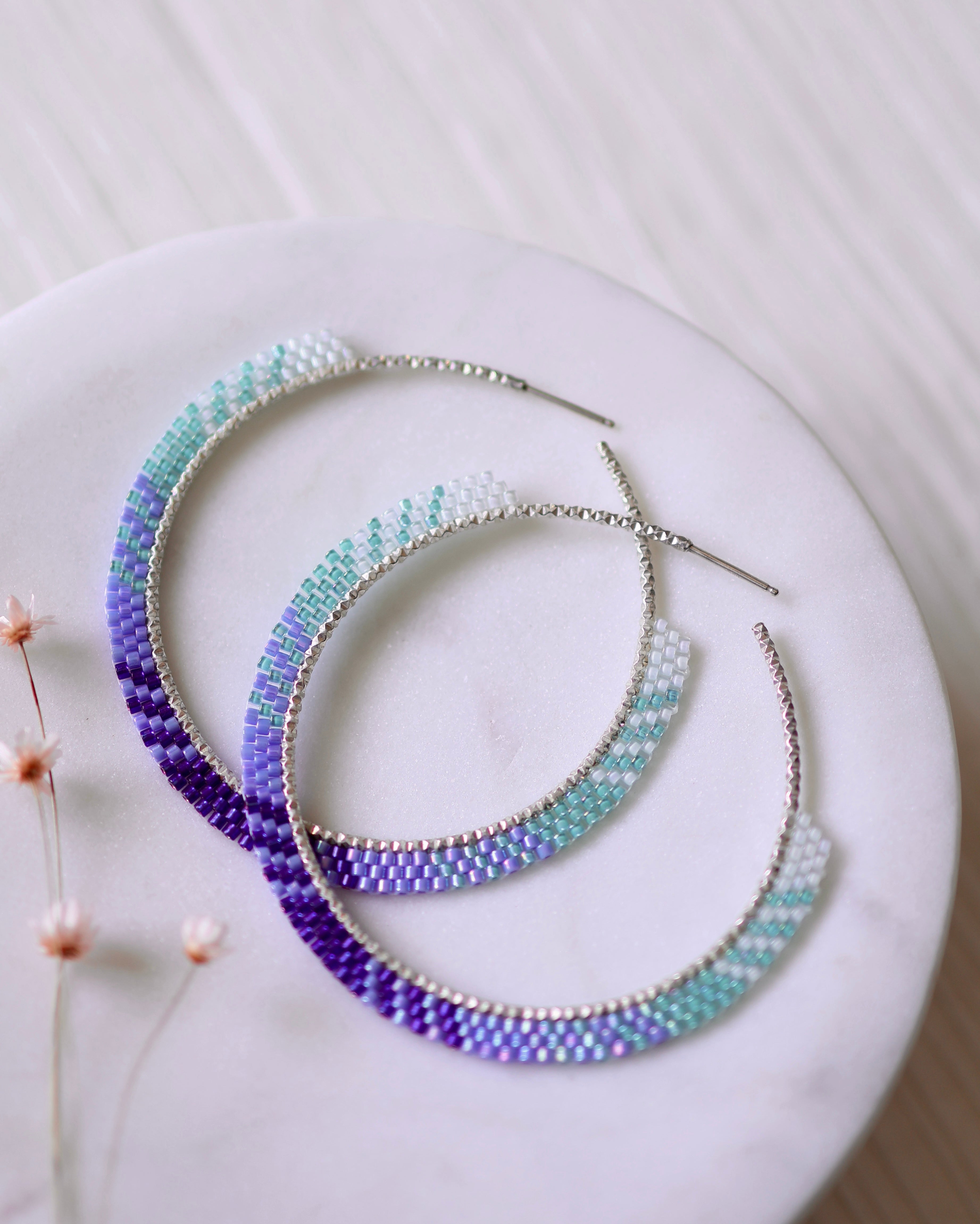 The Coastal Breeze Hoops by Lilac Sky Studio are beaded earrings featuring an ombré of white, turquoise, and purple Japanese glass seed beads, displayed on a round white dish beside small dried flowers.