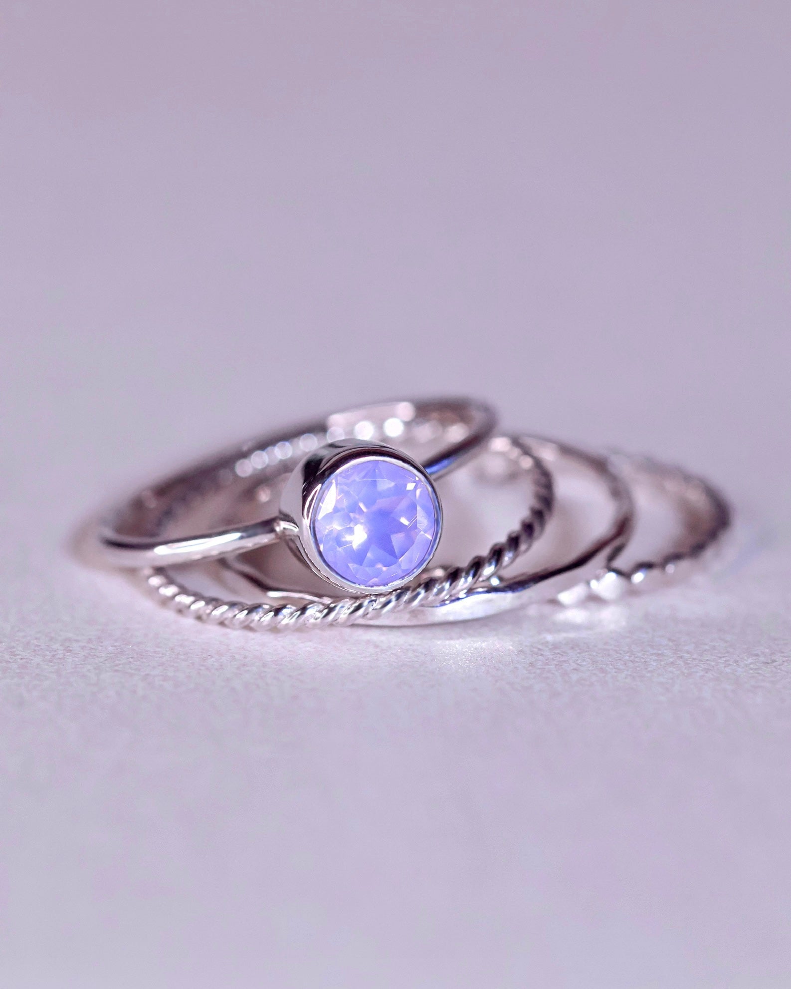 The Lavender Moon Ring Stacker Set by Lilac Sky Studio features sterling silver rings with a round, faceted Lavender Moon quartz, beautifully arranged on a light surface and paired with a gentle polishing cloth for lasting shine.
