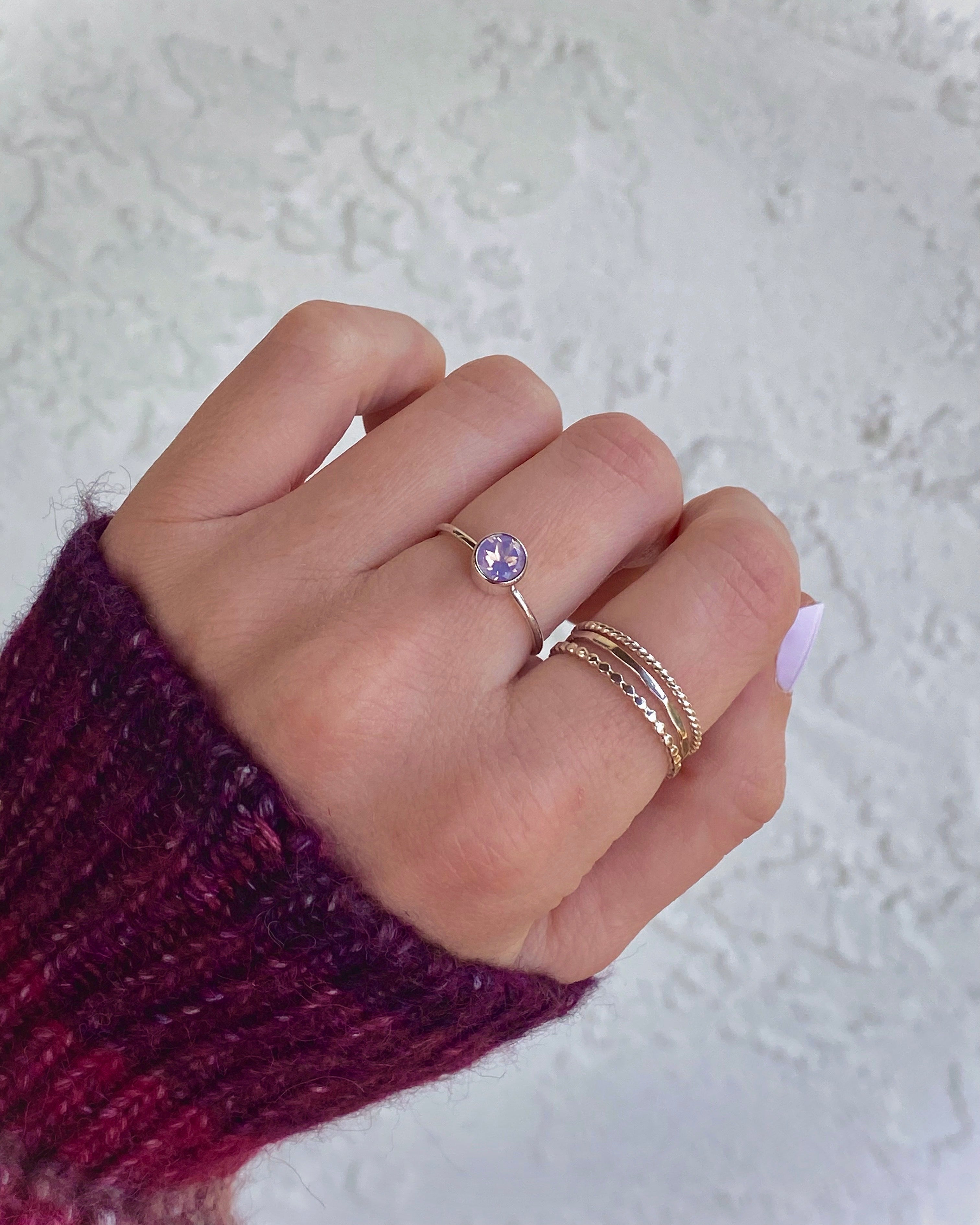 A hand with light pink nails wears three delicate rings from Lilac Sky Studio’s Lavender Moon Ring Stacker Set, set in sterling silver, against a textured white background. The wearer sports a purple knit sleeve.