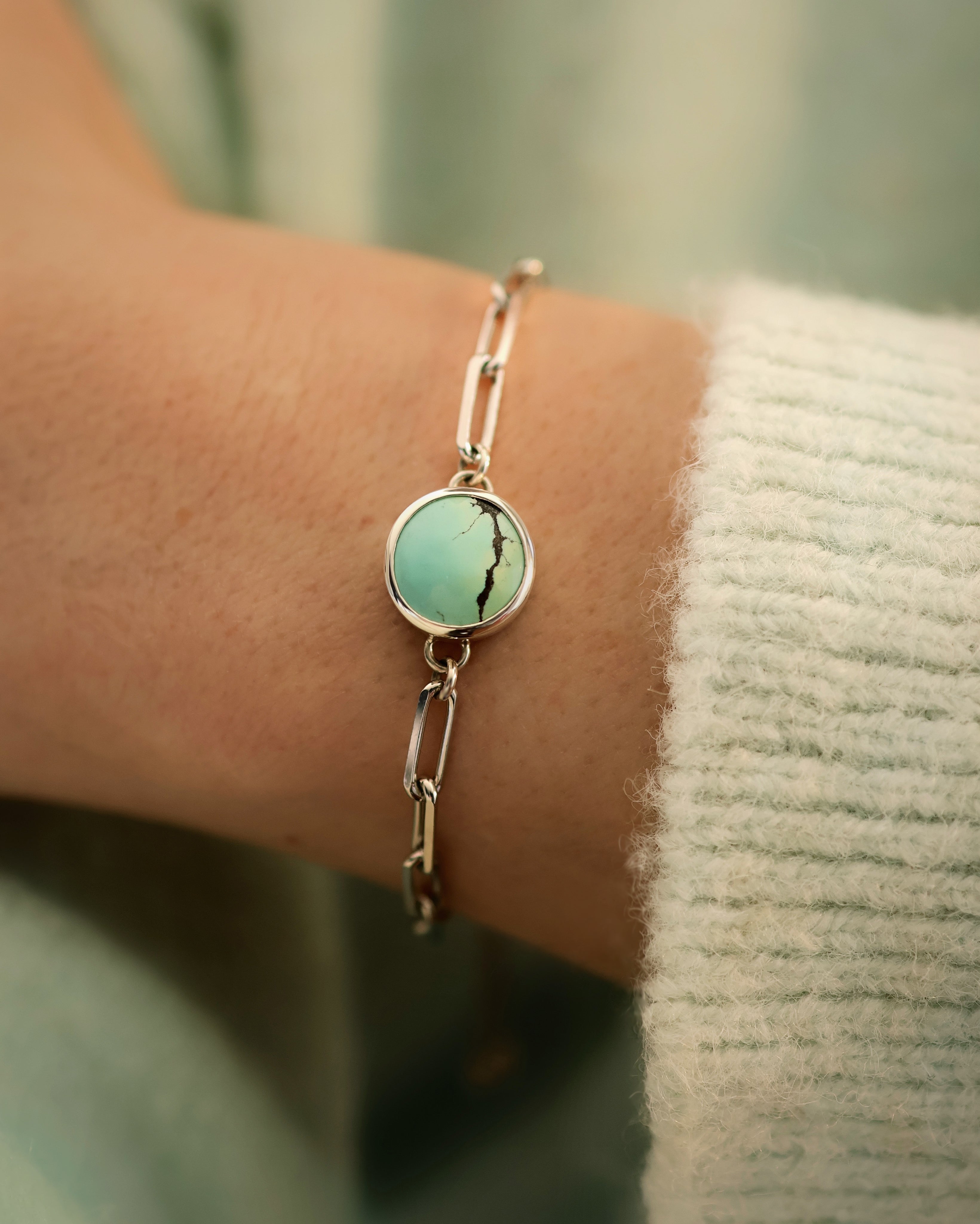 A close-up of the Lilac Sky Studio Treasure Mountain Turquoise Bracelet No. 2, featuring a round, light green turquoise stone with a dark vein set in sterling silver, worn on a wrist against a light knit sweater.