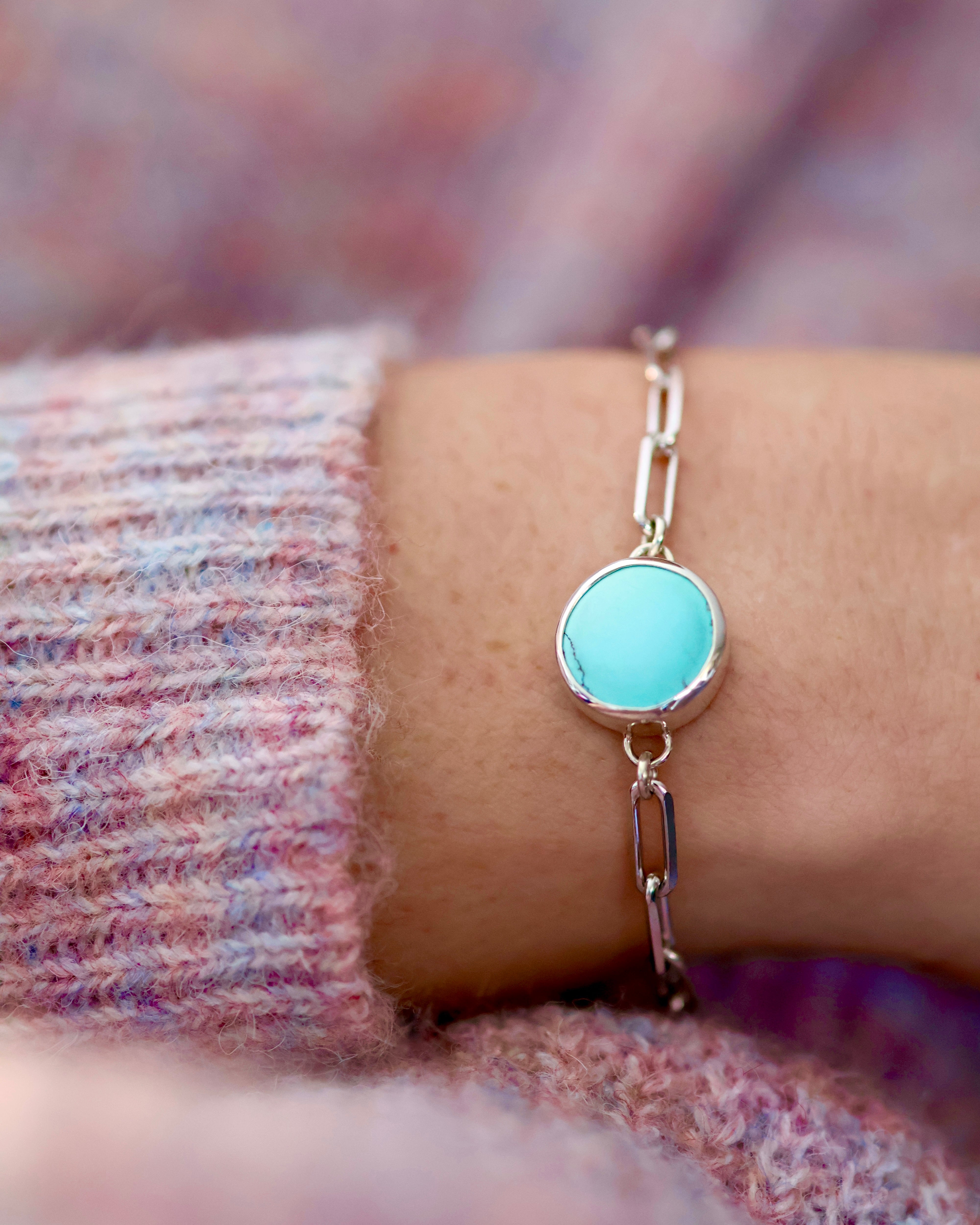 The Cloud Mountain Turquoise Bracelet No. 3 by Lilac Sky Studio, featuring a round turquoise stone on a silver chain, is shown on a wrist with a pink and purple textured sweater in the background.