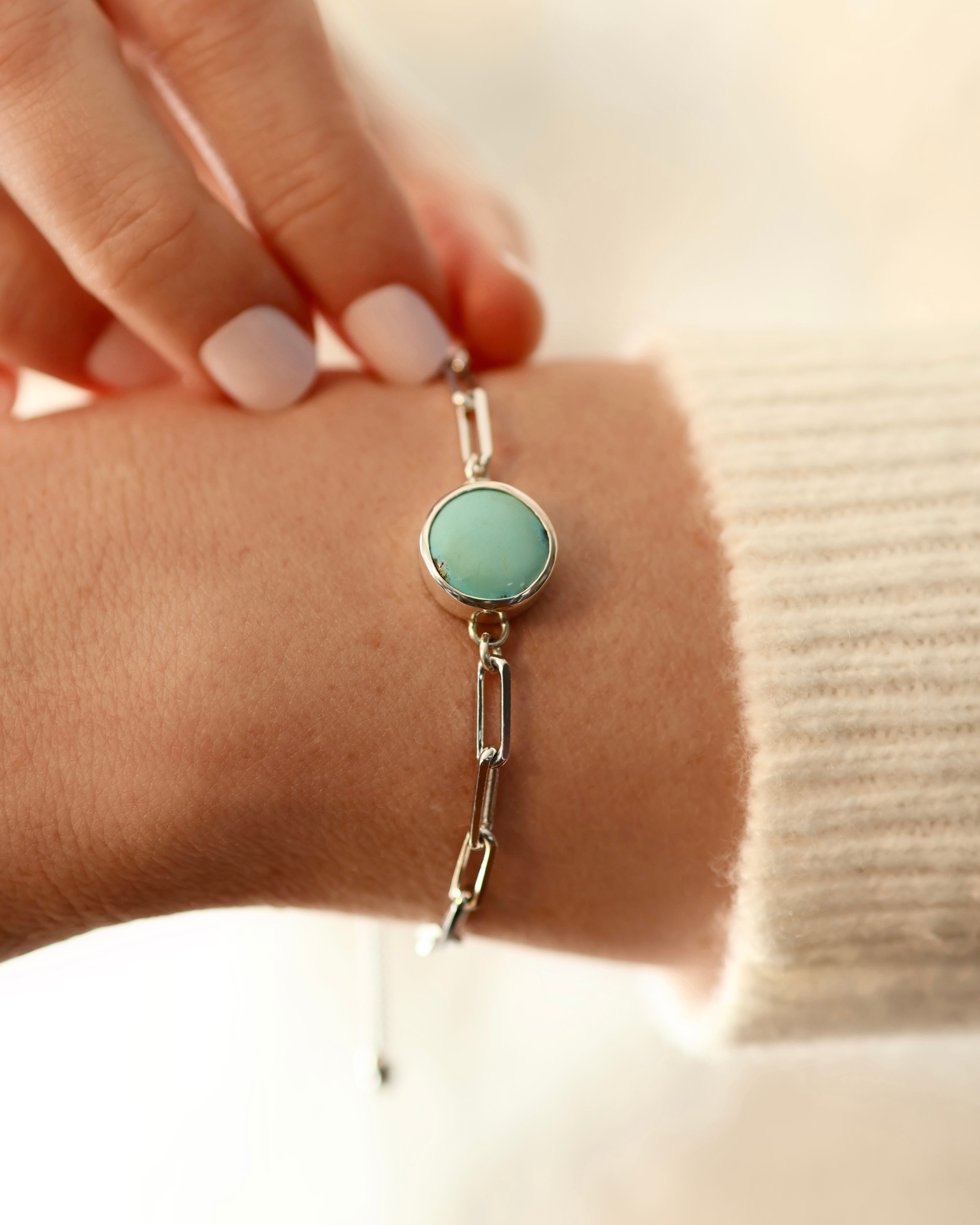 A hand wears Lilac Sky Studio's Treasure Mountain Turquoise Bracelet No. 1, featuring a round turquoise stone and adjustable sterling silver cable chain, shown against a cream-colored sweater.
