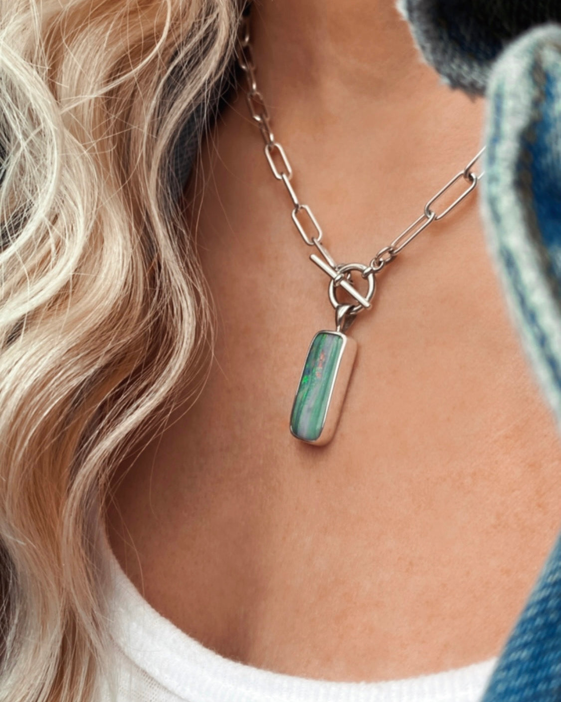 Close-up of a person wearing a handcrafted silver chain necklace with a rectangular green and pink stone pendant, white top, and light curly hair visible.