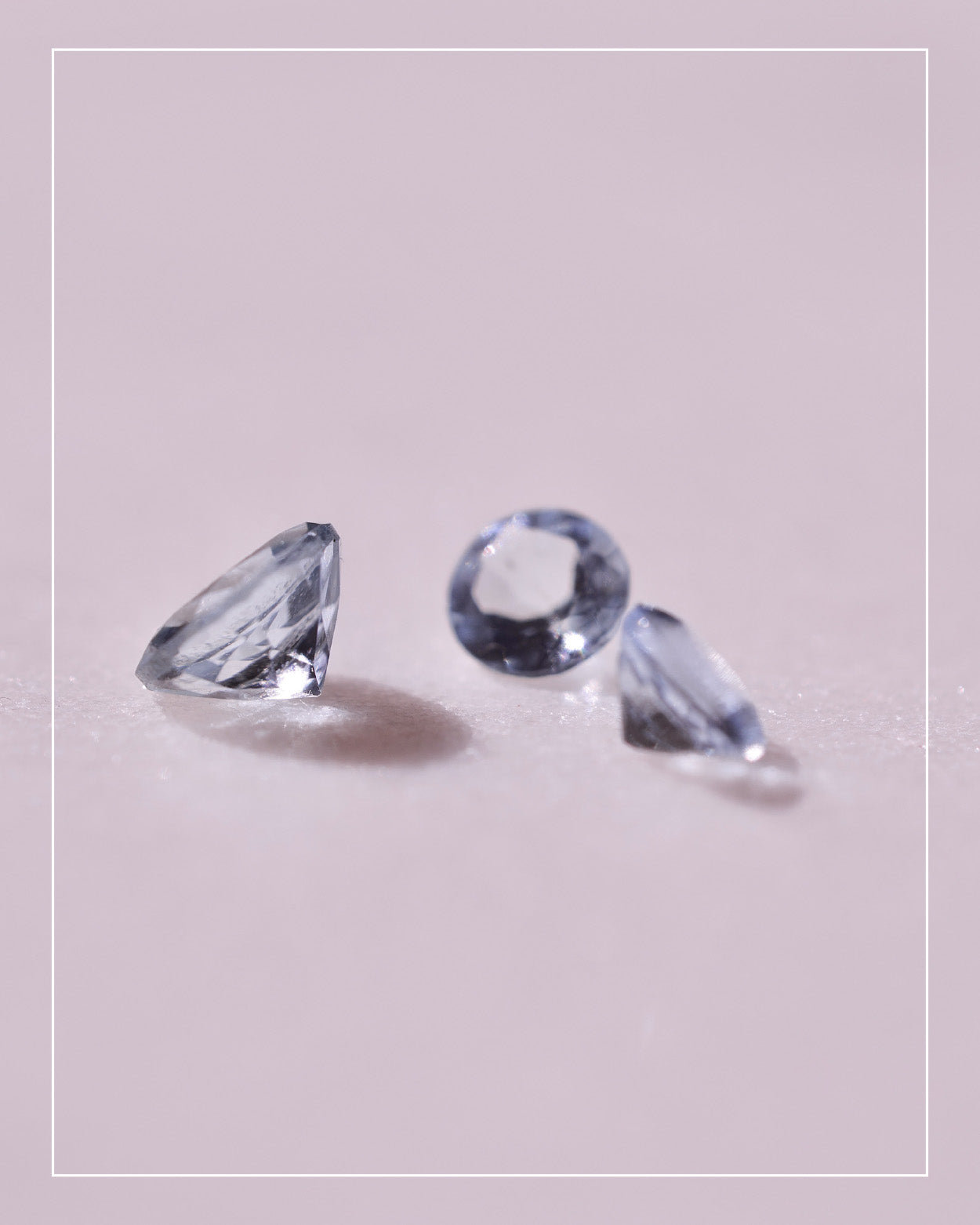 Three small, faceted blue gemstones—ethically sourced—are displayed on a light pink surface, arranged loosely with visible shadows beneath each stone.