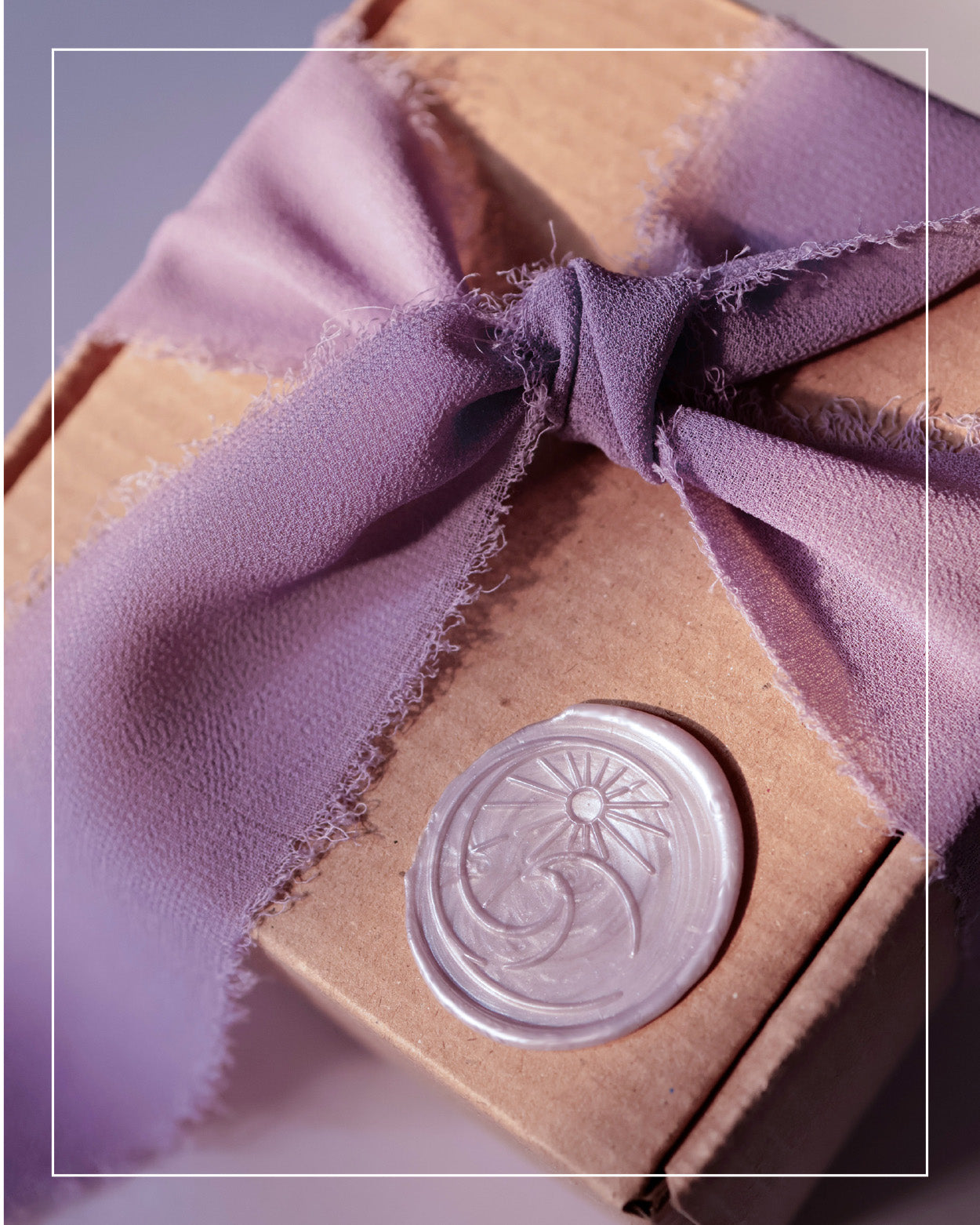 A cardboard box wrapped with a frayed lavender ribbon, featuring a decorative wax seal with a sun and crescent moon design, perfect for presenting ethically sourced gold jewelry.