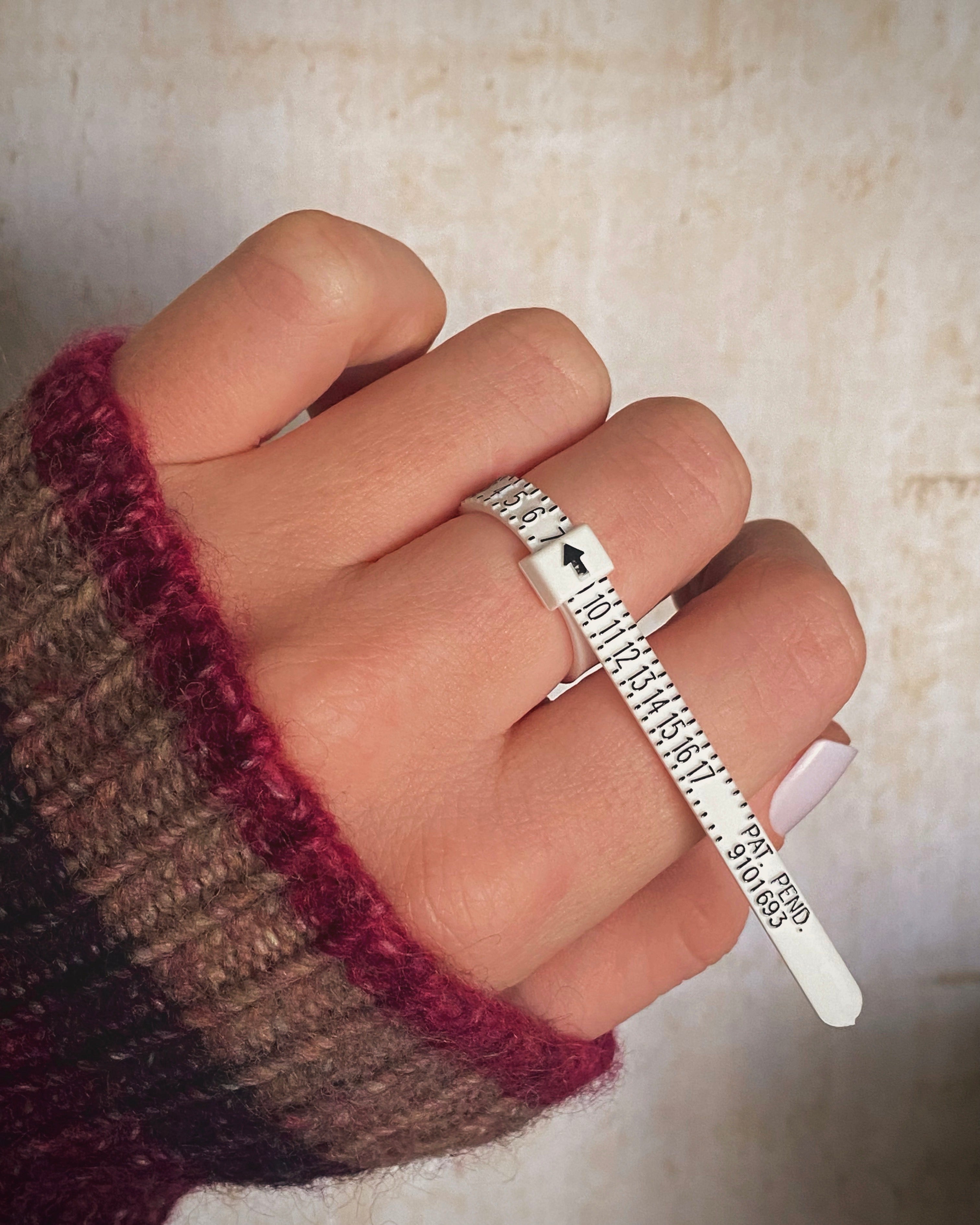 A hand wearing a striped sweater uses a white plastic ring sizer to measure finger circumference, with the arrow pointing to the ring size—perfect for selecting beautifully handcrafted jewelry.