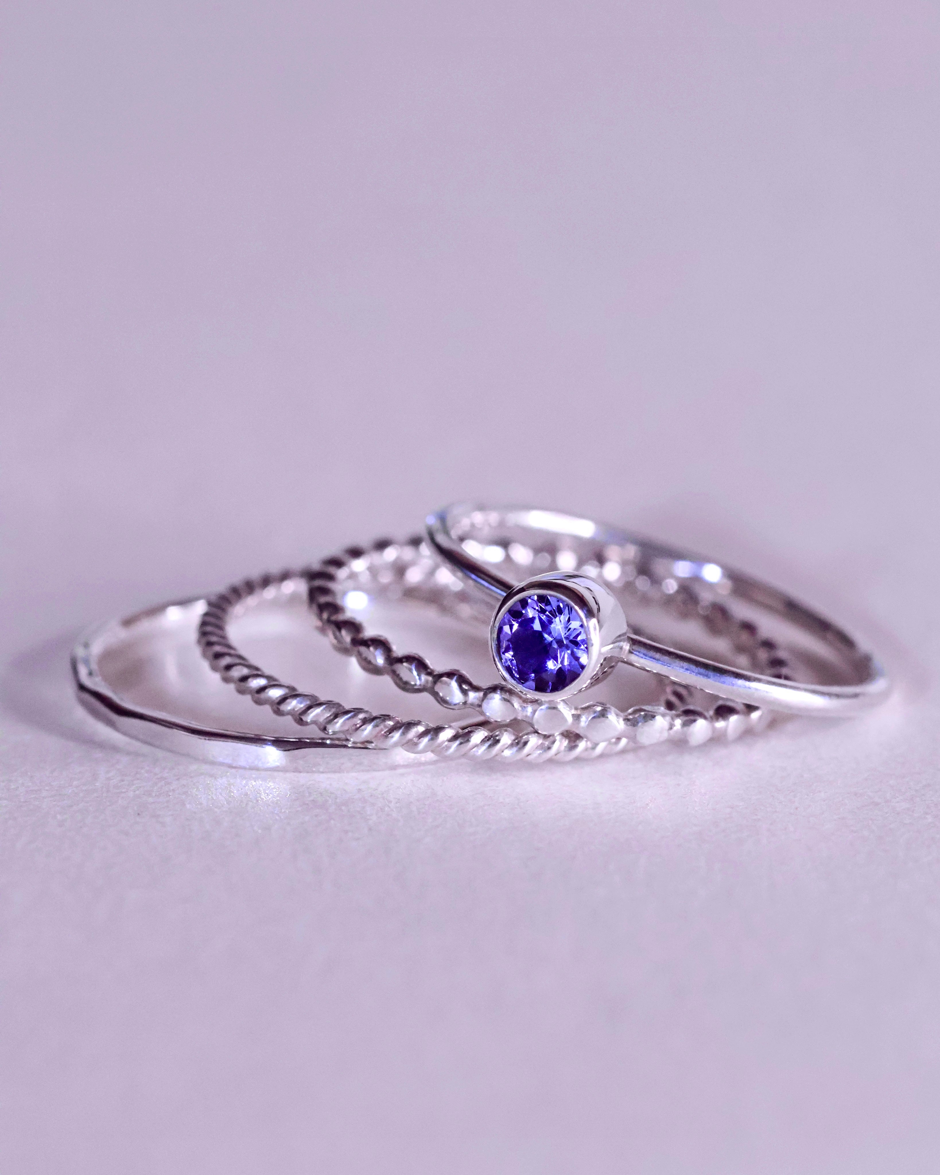 The Tanzanite Ring Stacker Set by Lilac Sky Studio features four sterling silver rings—one with a round Tanzanite gemstone and three with unique band designs—perfect for anyone who loves distinctive gemstone jewelry.