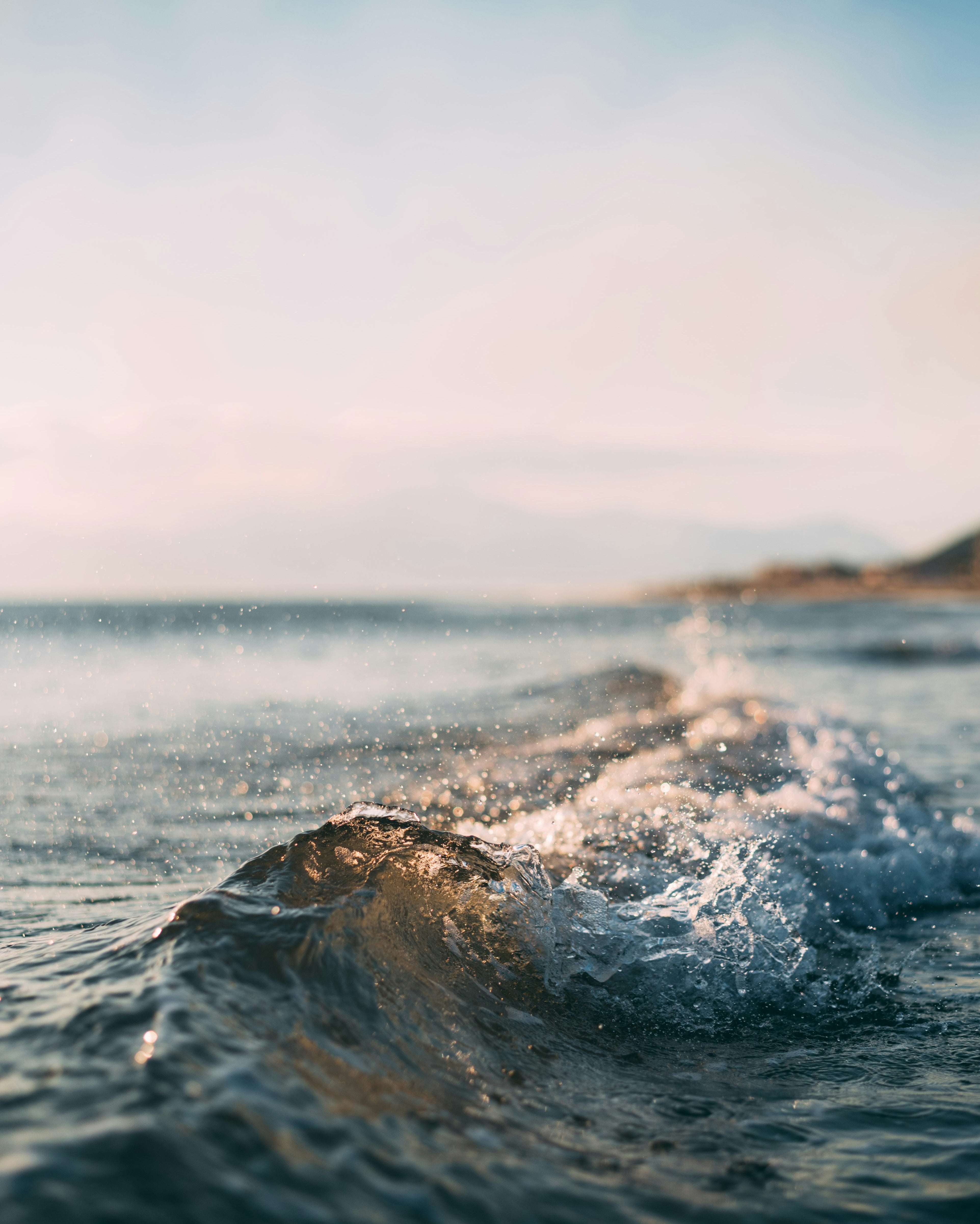 Close-up of a small ocean wave breaking near the shore with distant mountains in the background under a soft, pastel sky, resembling the smooth curves of handcrafted jewelry.