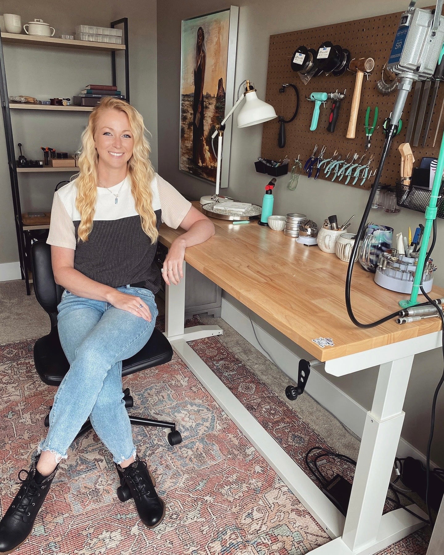A woman sits on a chair next to a wooden workbench with tools hanging on a pegboard in a well-organized workspace, carefully crafting handcrafted gold and gemstone pieces.