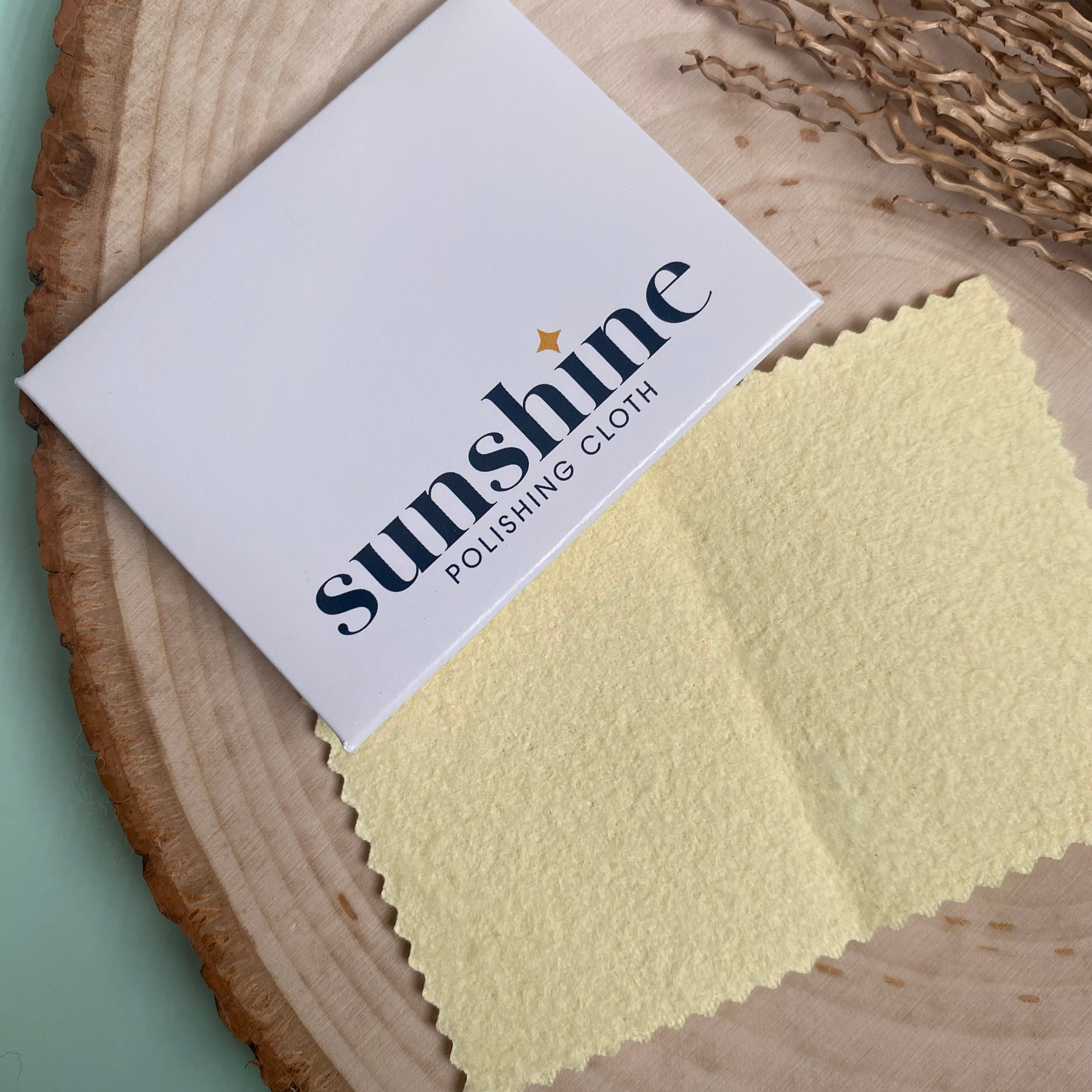 A yellow polishing cloth for jewelry is partially pulled out from a white envelope labeled "Sunshine Polishing Cloth," placed on a wooden surface. Ideal for keeping your handcrafted pieces and gemstone treasures shining bright.