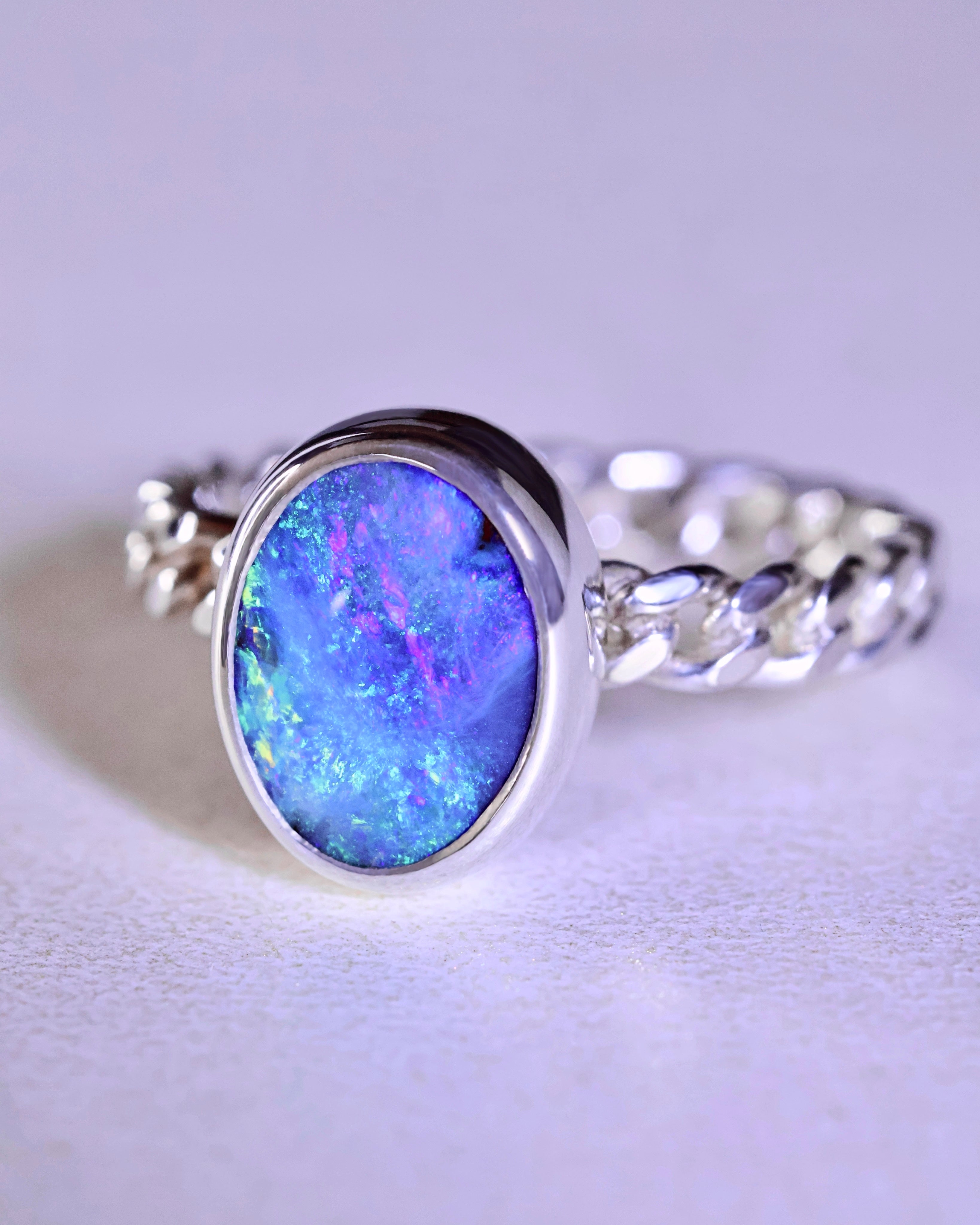 The Boulder Opal Ring by Lilac Sky Studio features a large oval opal in blue, purple, and green hues set in sterling silver, displayed on a light surface.