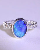 The Boulder Opal Ring by Lilac Sky Studio is a sterling silver band with a braided design, showcasing an oval, gem-quality opal gemstone with vibrant blue, green, and purple hues.