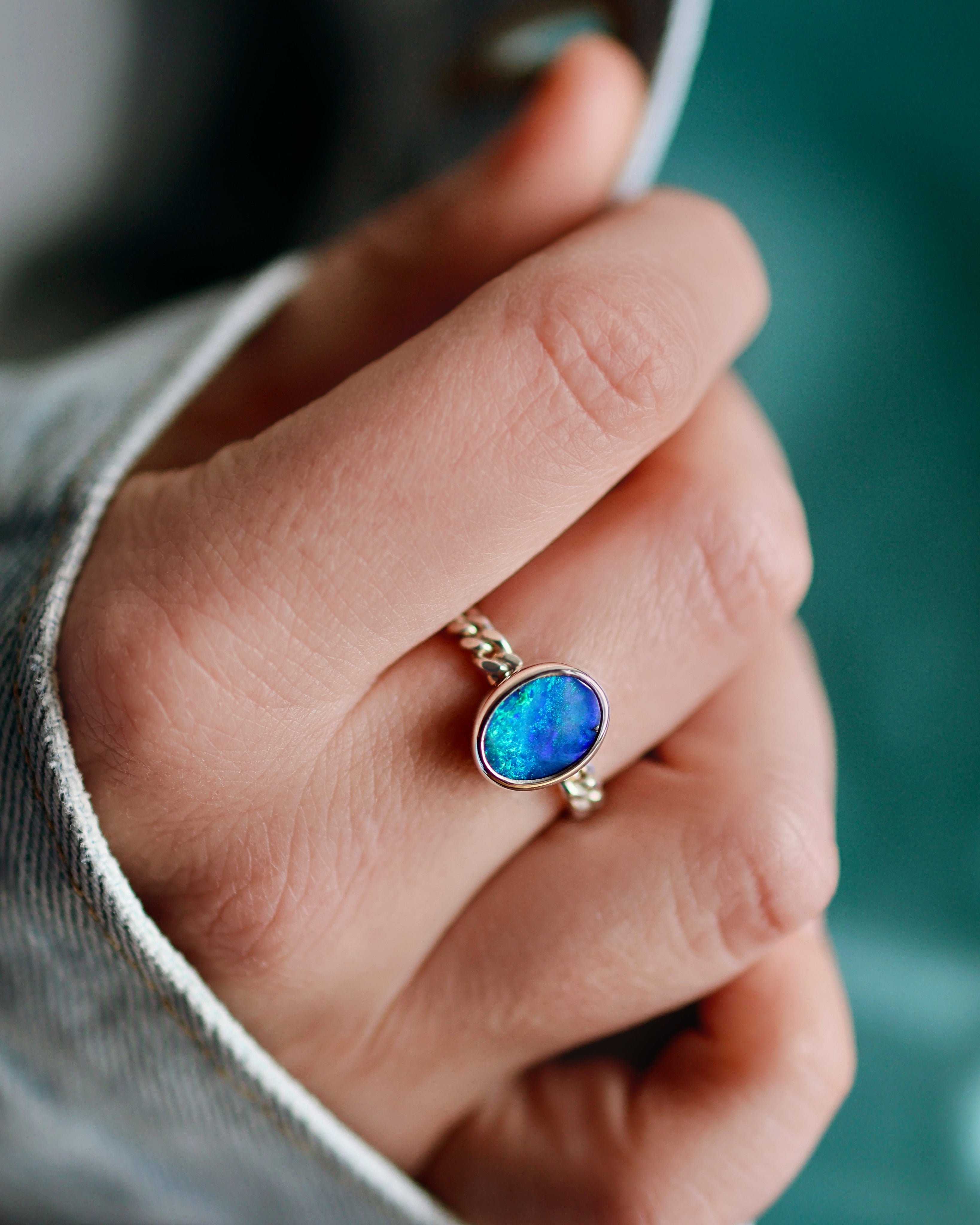 A hand adorned with Lilac Sky Studio’s Boulder Opal Ring, featuring a sterling silver band and an oval-shaped blue-green opal, is partially covered by a light denim sleeve.