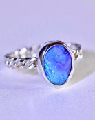 The Boulder Opal Ring by Lilac Sky Studio showcases a gem-quality oval opal in a bezel setting on a twisted sterling silver band, displayed on a light surface.