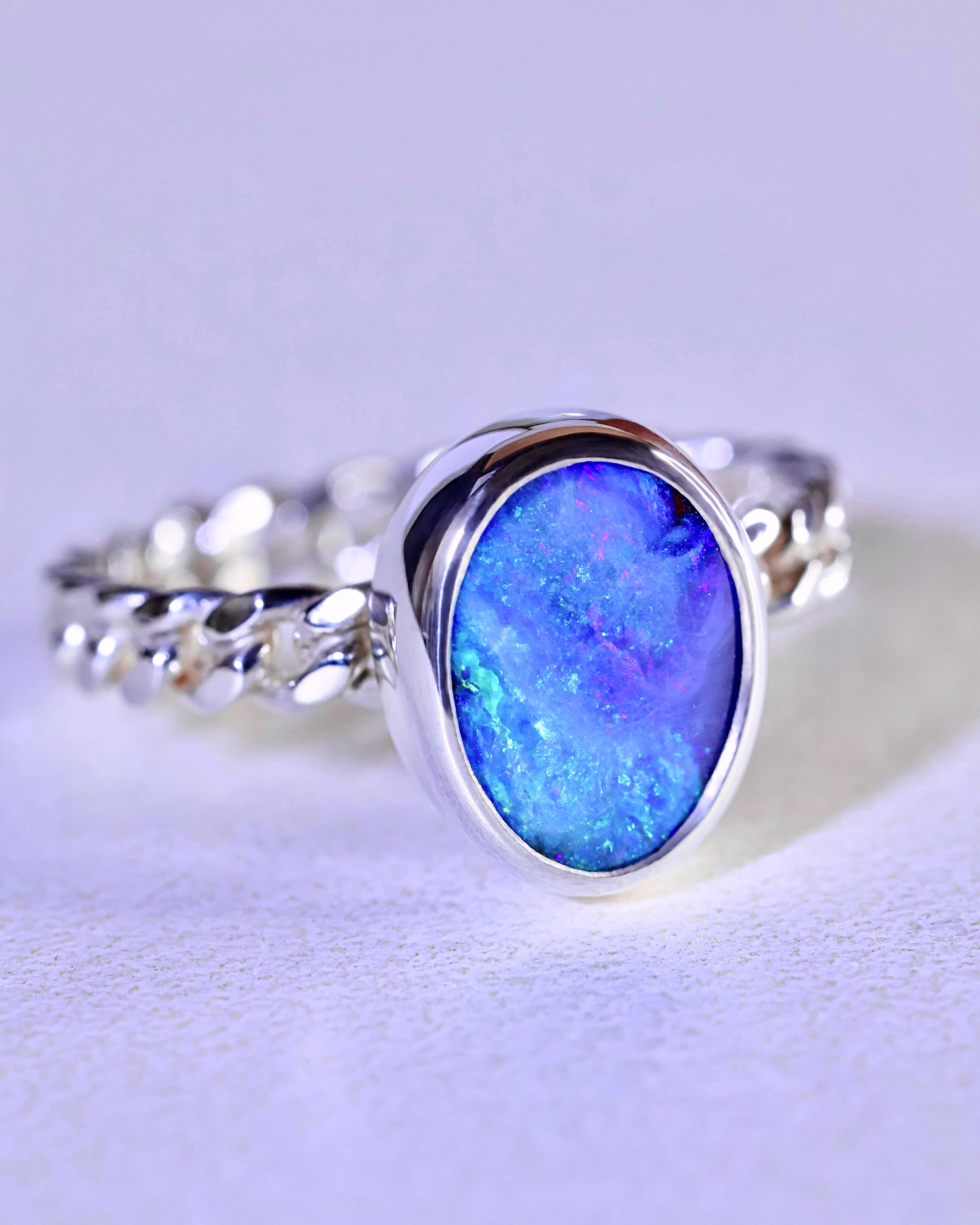 The Boulder Opal Ring by Lilac Sky Studio showcases a gem-quality oval opal in a bezel setting on a twisted sterling silver band, displayed on a light surface.