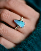 A hand wearing an ethically sourced silver ring with a blue-green opal stone, shown against a teal fabric background—an elegant piece of jewelry.
