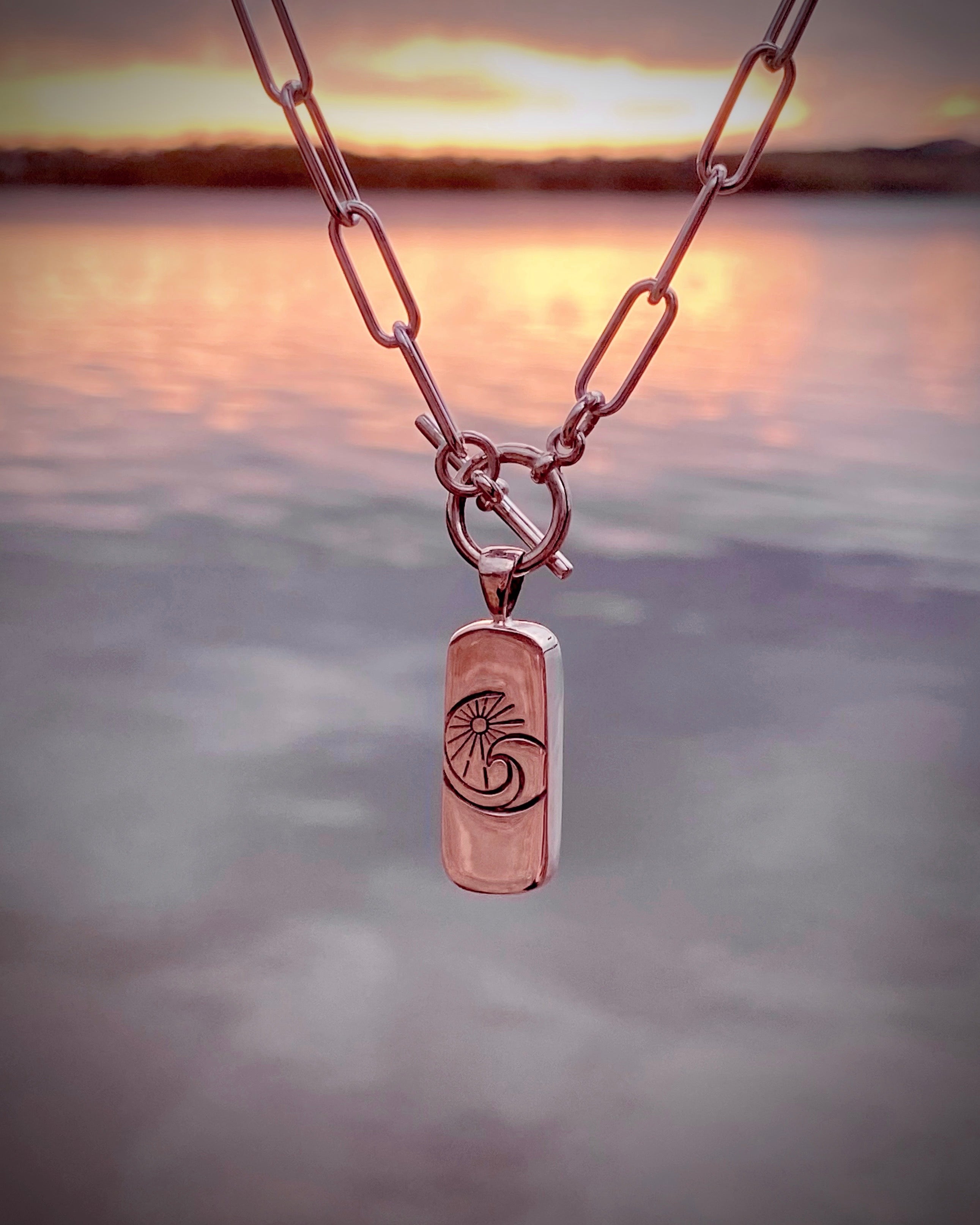 The Boulder Opal Necklace by Lilac Sky Studio features a shimmering opal pendant on a sleek paperclip chain, elegantly displayed against a blurred sunset over water.