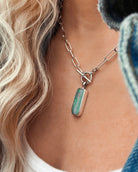 A close-up of a person wearing Lilac Sky Studio's Boulder Opal Necklace, featuring a sterling silver paperclip chain and a rectangular green-blue natural boulder opal pendant, against light skin and wavy blonde hair.
