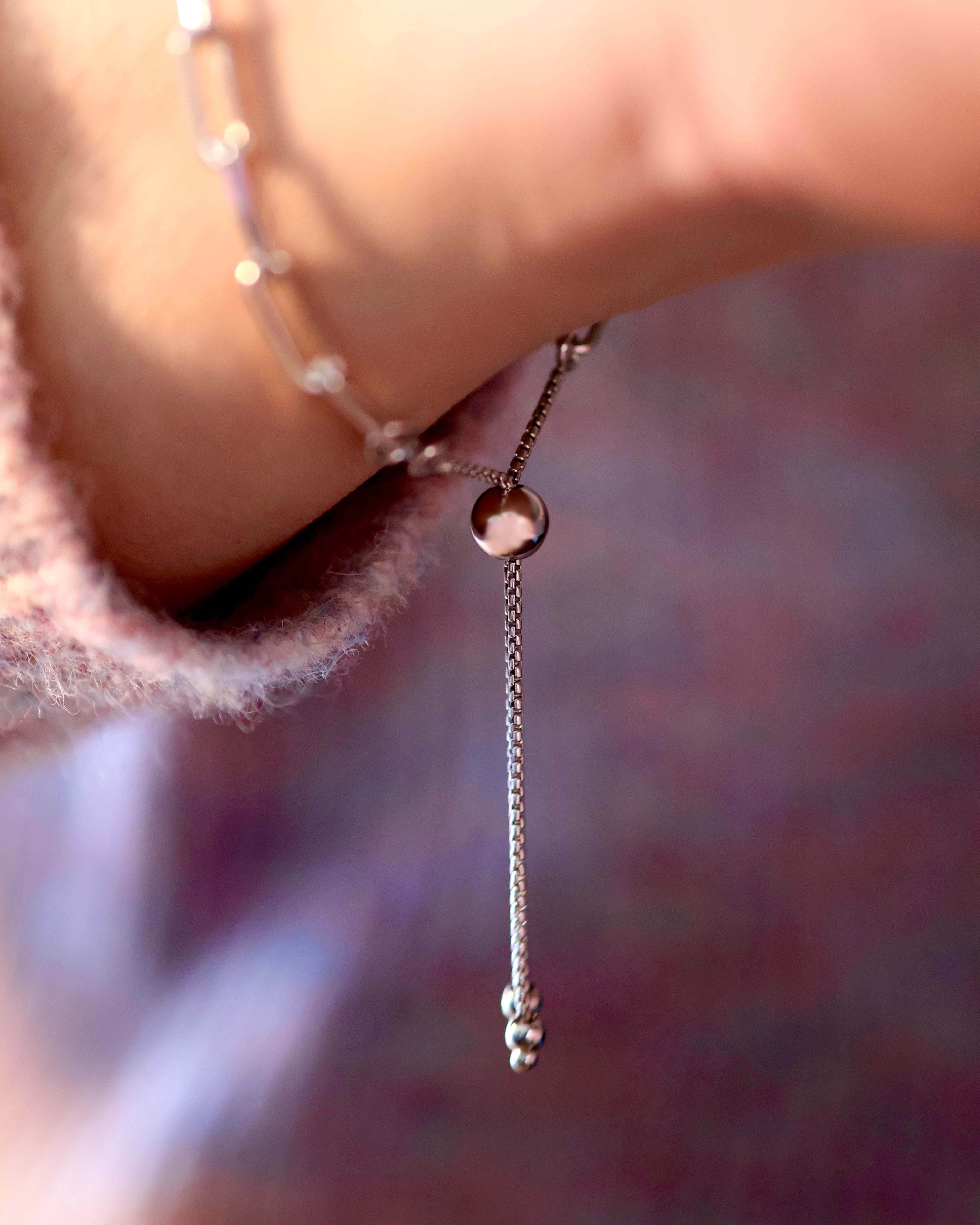 Close-up of a wrist wearing the Cloud Mountain Turquoise Bracelet No. 3 by Lilac Sky Studio, featuring an adjustable sterling silver cable chain with a dangling end, set against a blurred purple and pink background.