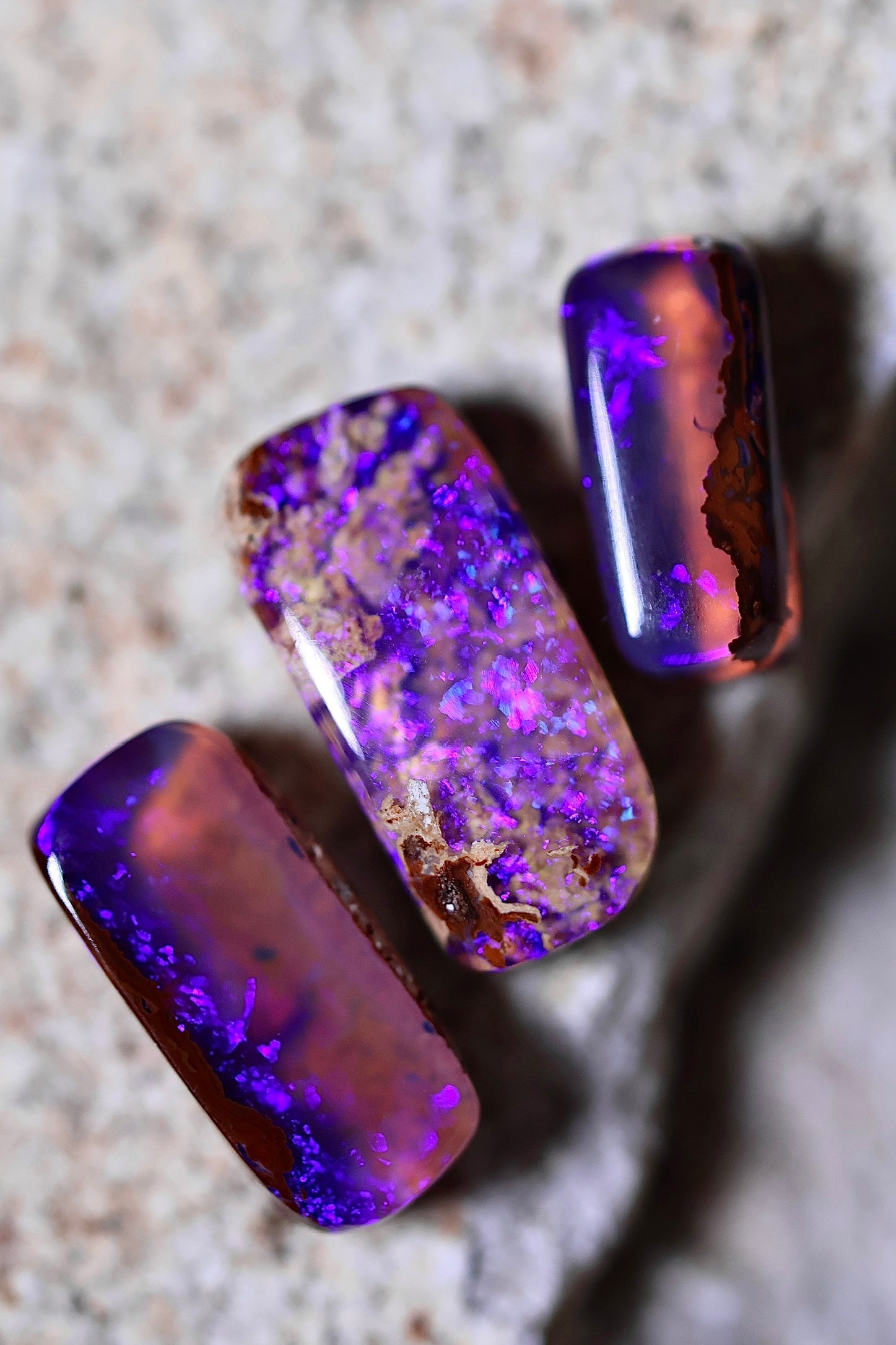 Three polished opal stones with purple and blue iridescent patterns are displayed on a textured stone surface, perfect for ethically sourced jewelry or to accent gold settings.