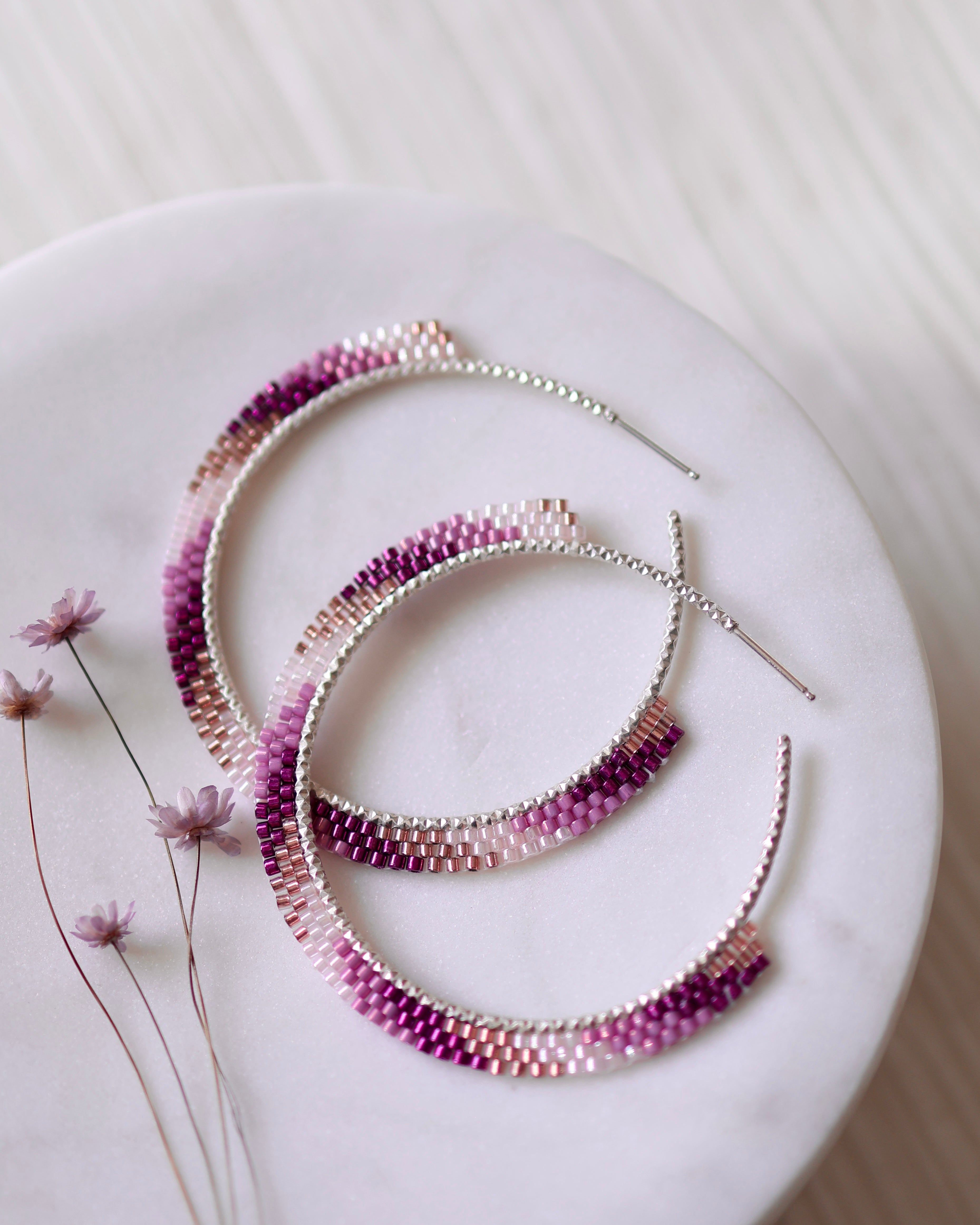The Sunkissed Hoops by Lilac Sky Studio, handwoven with Japanese glass seed beads in a pink-to-purple gradient, are displayed on a round white marble surface beside dried flowers.