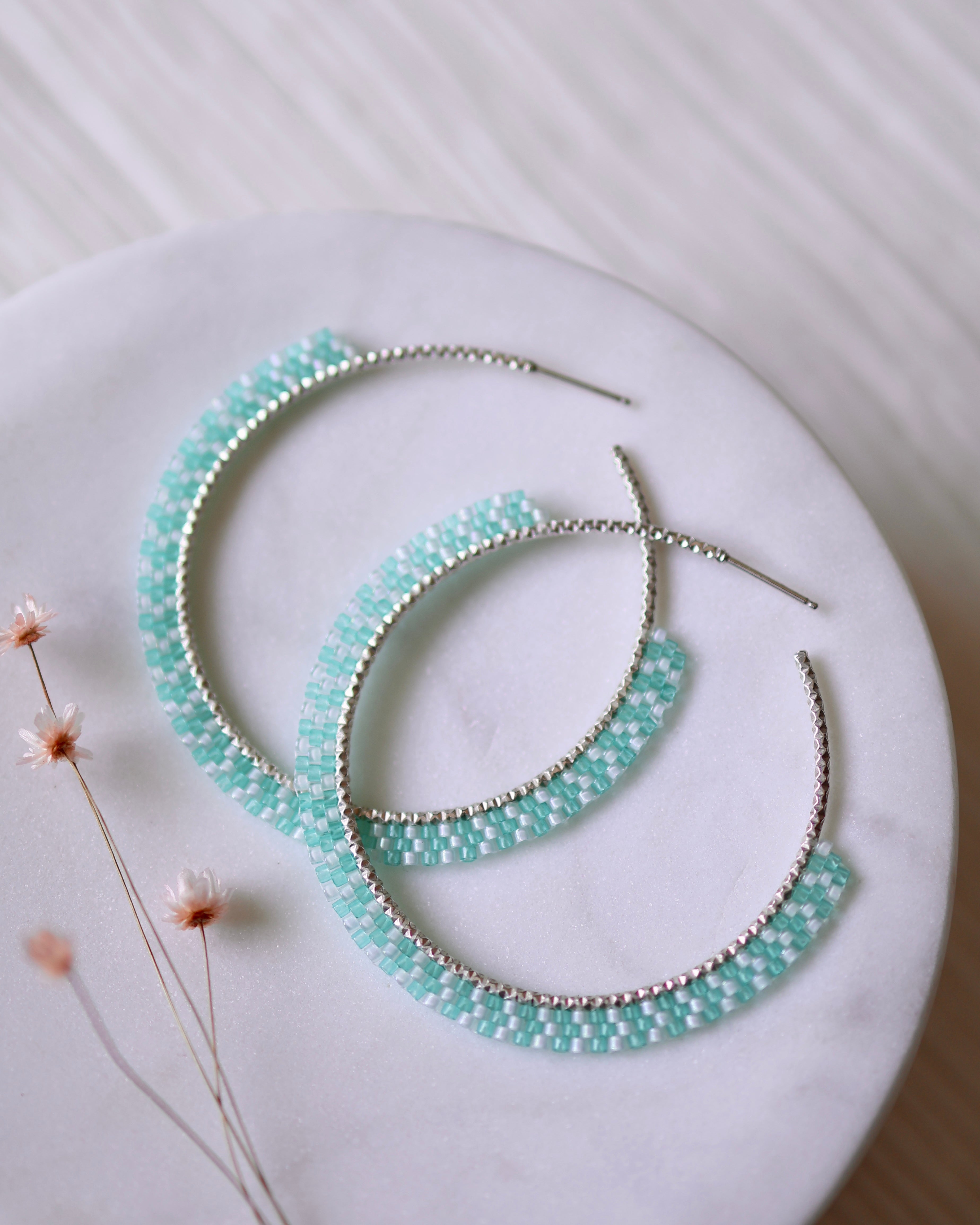 The Party Cove Hoops by Lilac Sky Studio are large sterling silver hoop earrings with light blue and white Japanese glass seed beads, shown on a round white marble surface beside small dried flowers.