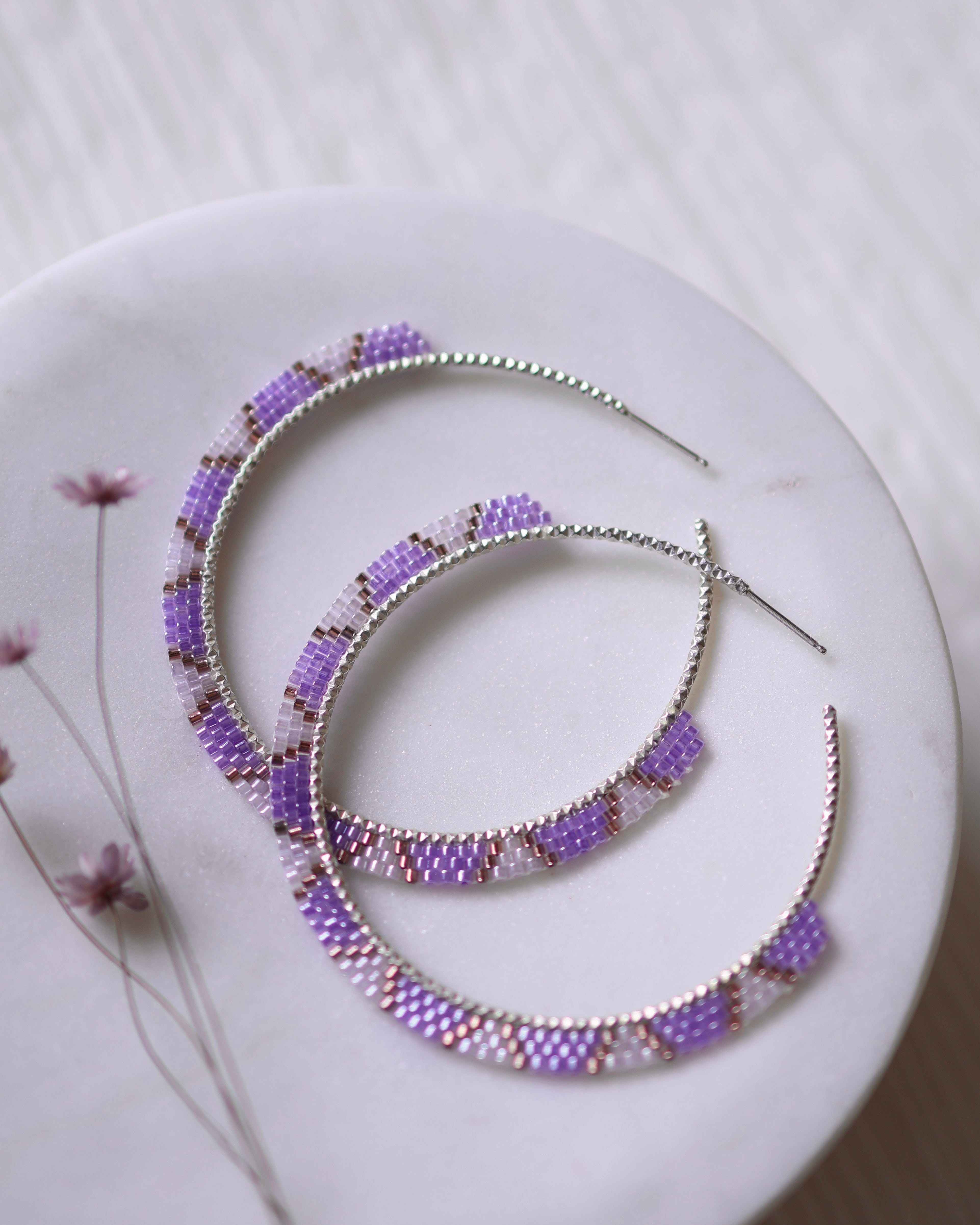 Lavender Rose Hoops by Lilac Sky Studio—two sterling silver earrings adorned with purple, lilac, and light pink Japanese glass seed beads—are displayed on a white round surface with delicate dried flowers beside them.