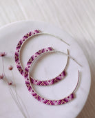 The Sunset Glow Hoops by Lilac Sky Studio are large curved handwoven earrings featuring a pink and purple beaded pattern with Japanese glass seed beads, displayed on a round white surface accented by small dried flowers.