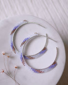 The Satin Rose Hoops by Lilac Sky Studio are handwoven earrings crafted with purple, white, and rose gold Japanese glass beads on sterling silver hoops, shown on a round white surface beside small dried flowers.