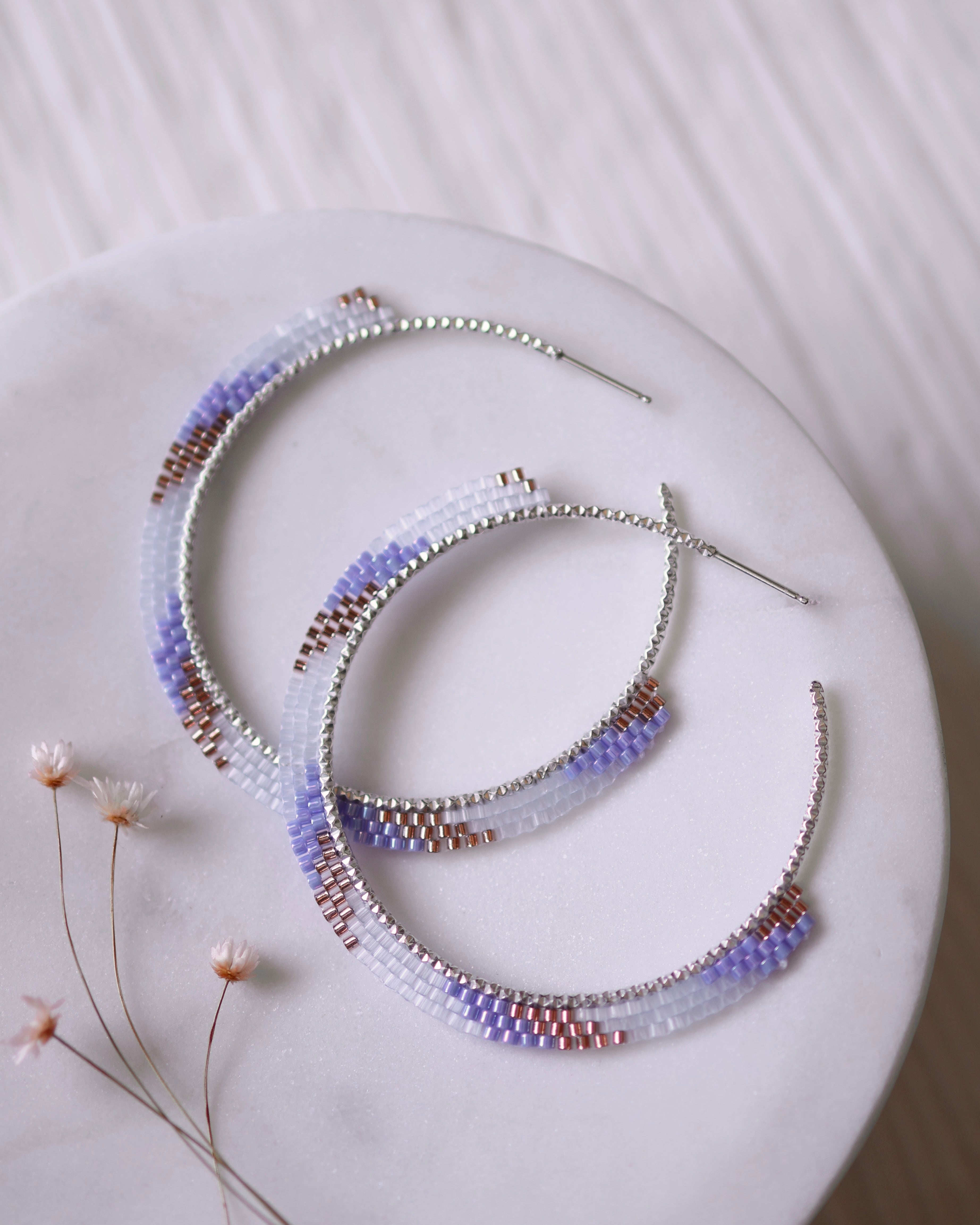 The Satin Rose Hoops by Lilac Sky Studio are handwoven earrings crafted with purple, white, and rose gold Japanese glass beads on sterling silver hoops, shown on a round white surface beside small dried flowers.