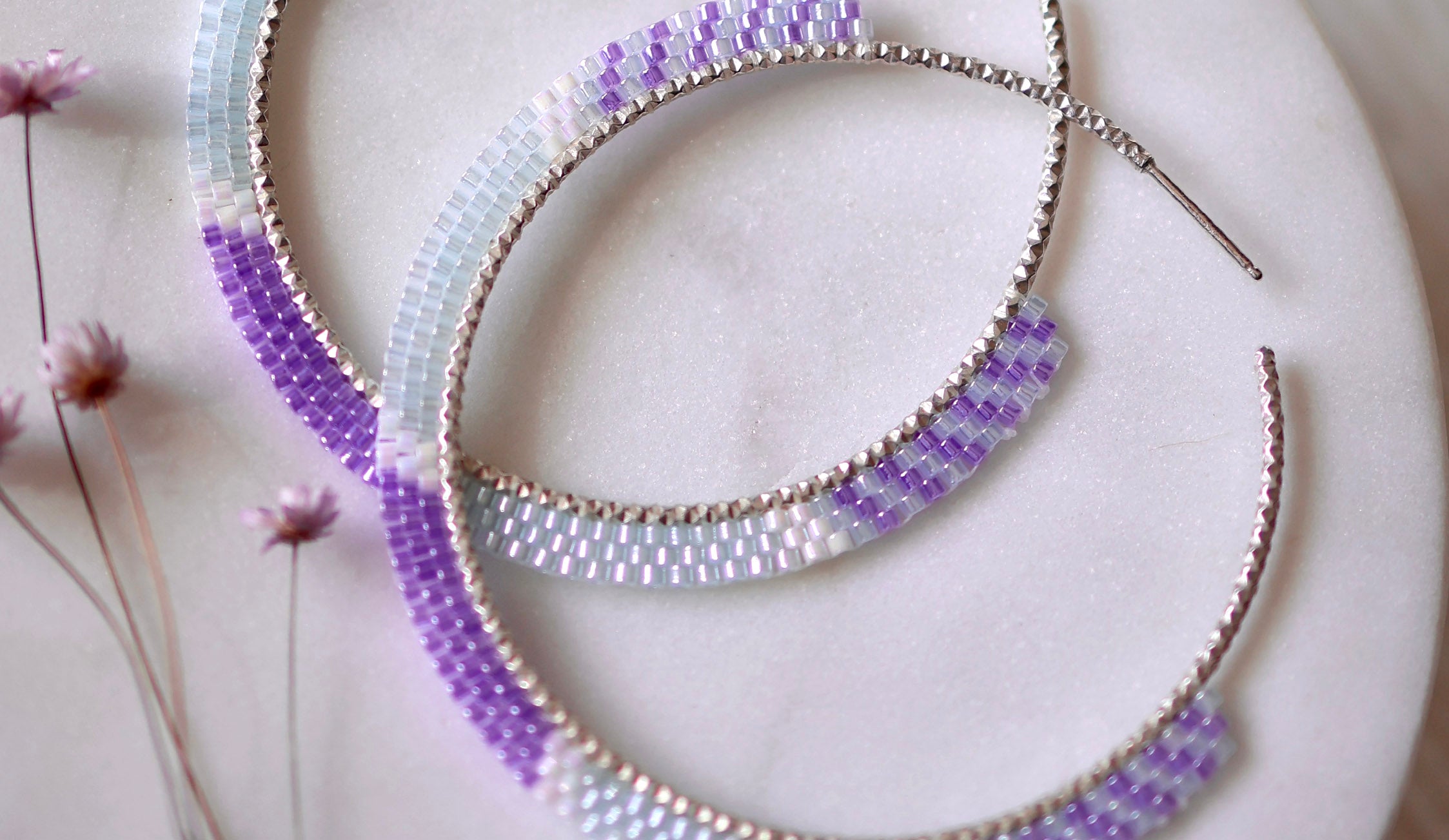 Two large hoop earrings with a beaded pattern in shades of purple and white lie on a marble surface next to small dried flowers, showcasing artisan jewelry crafted with ethically sourced materials.