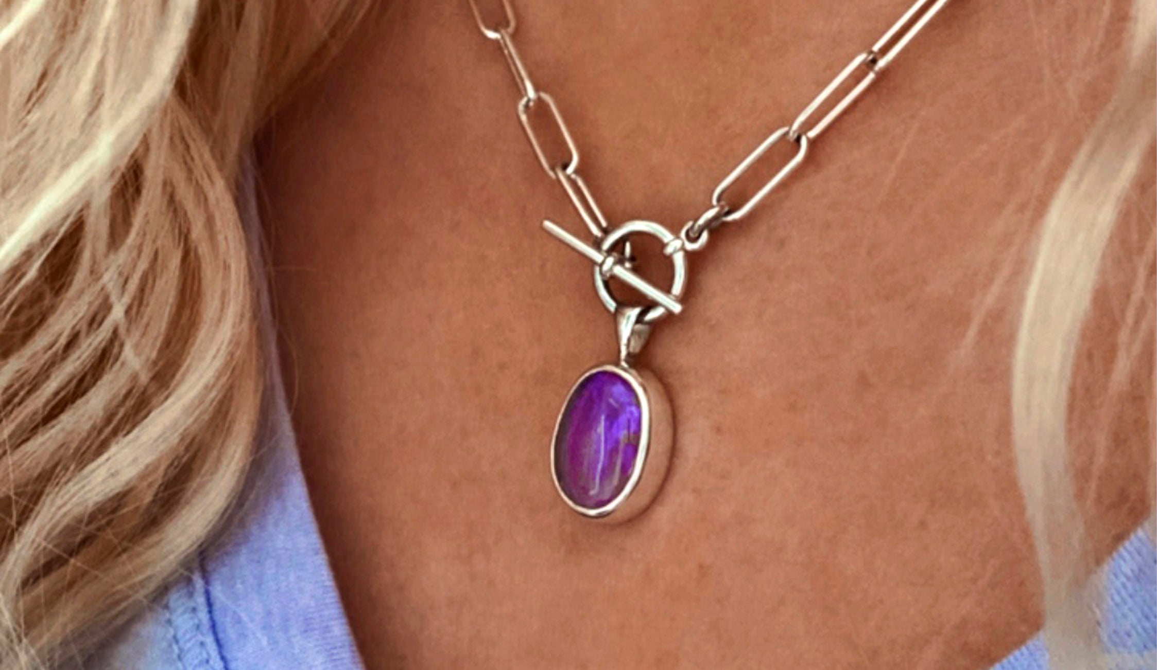 A close-up of a silver chain necklace with a toggle clasp and a purple oval gemstone pendant, showcasing stunning handcrafted jewelry resting on a person's chest.