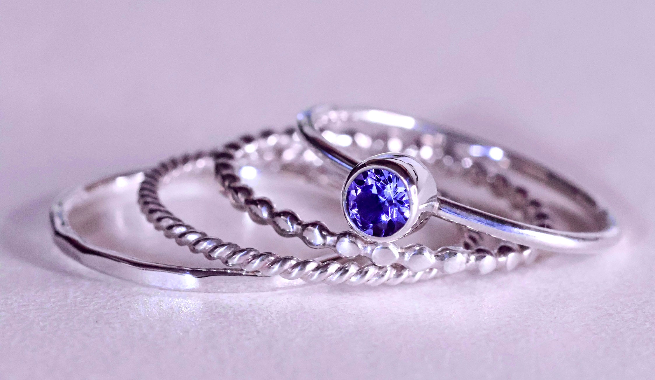 A set of four silver rings, including a plain band, a twisted band, a beaded band, and a ring featuring an ethically sourced round purple gemstone—beautiful additions to your jewelry collection.
