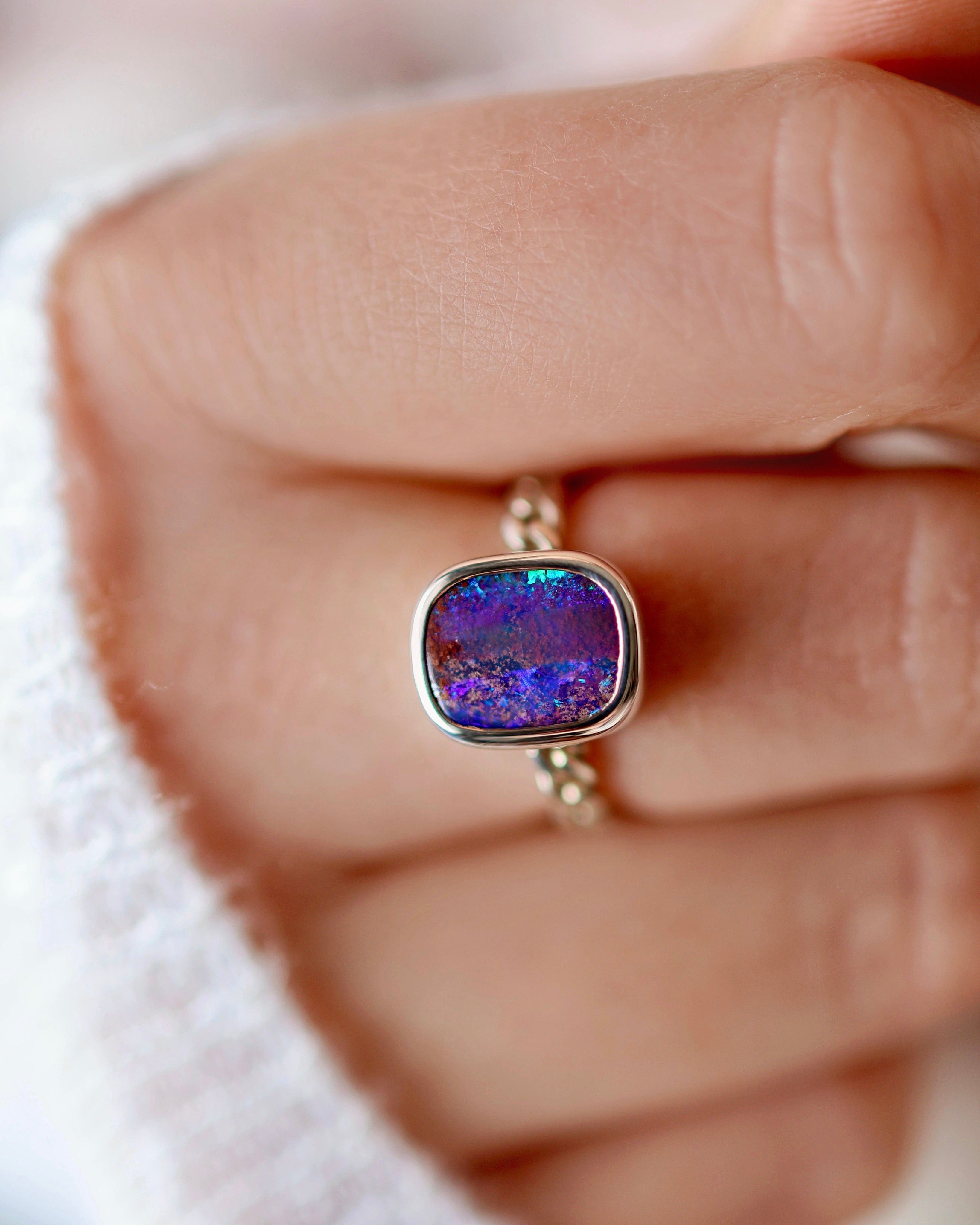 A close-up of a hand wearing a silver ring with an ethically sourced, rectangular gemstone featuring multicolored purple and blue tones.