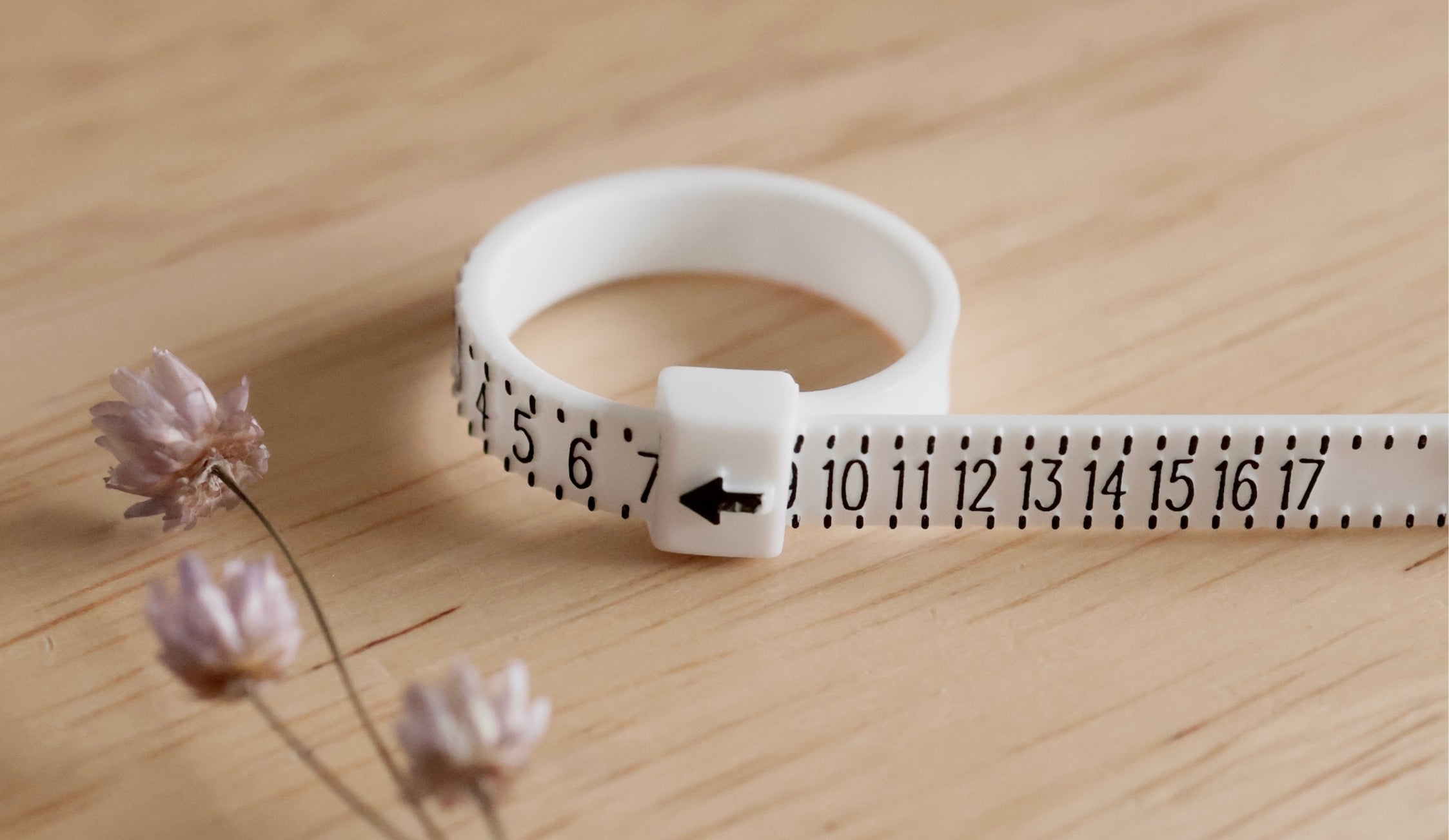 A white adjustable ring sizer with numbers and an arrow indicator, perfect for sizing handcrafted jewelry, is placed on a wooden surface next to small dried flowers.