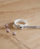 The Lilac Sky Studio Ring Sizer, a white adjustable tool with measurement markings, half-size increments, and an arrow, is displayed on a light wooden surface next to small dried flowers.