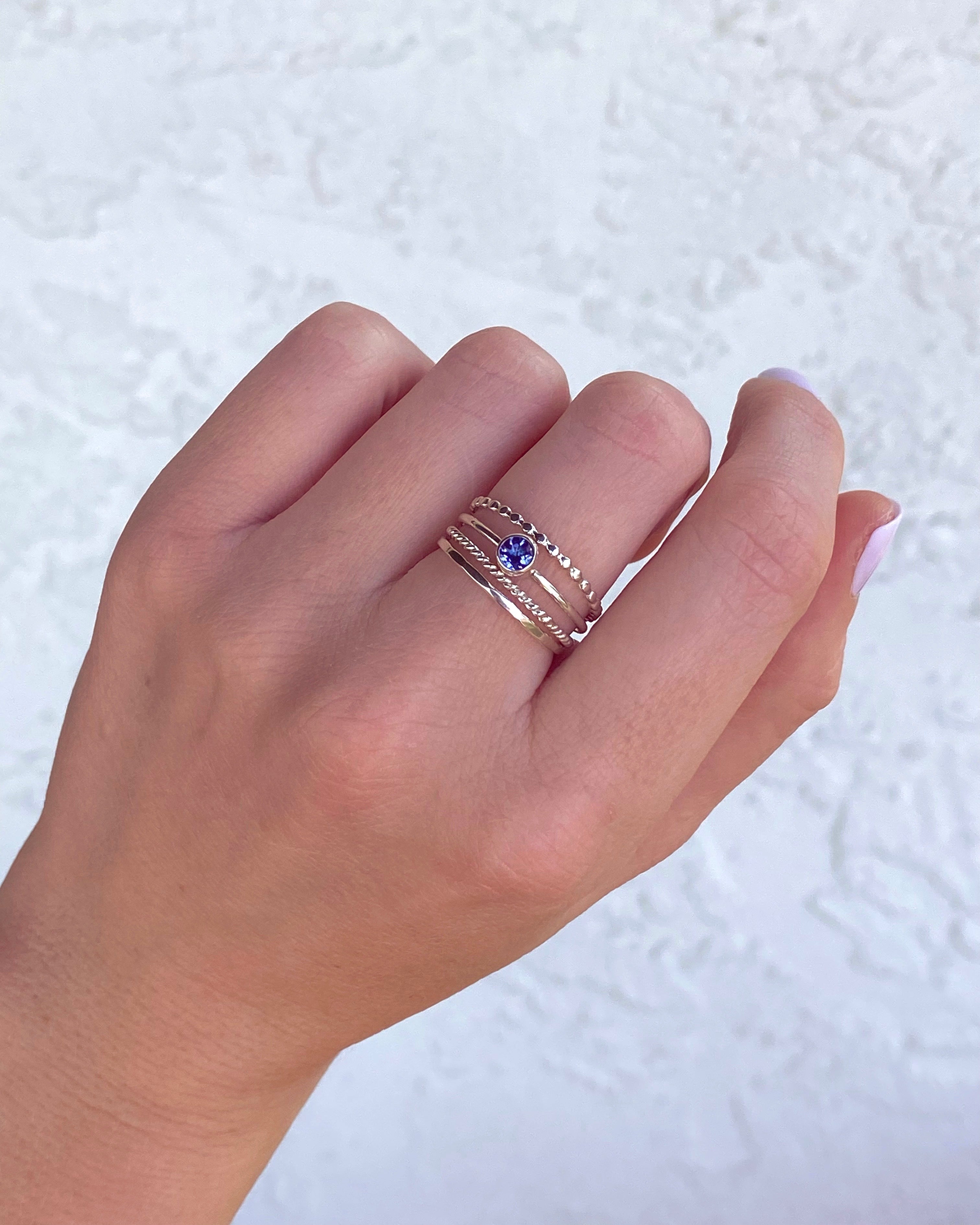 A hand with light skin tone wears the Lilac Sky Studio Tanzanite Ring Stacker Set—multiple thin sterling silver rings, including one with a small blue tanzanite gemstone—against a textured white background.