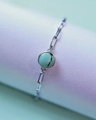 The Treasure Mountain Turquoise Bracelet No. 2 by Lilac Sky Studio is a sterling silver adjustable cable chain with a round turquoise stone featuring a dark vein, shown on a pastel-colored surface.