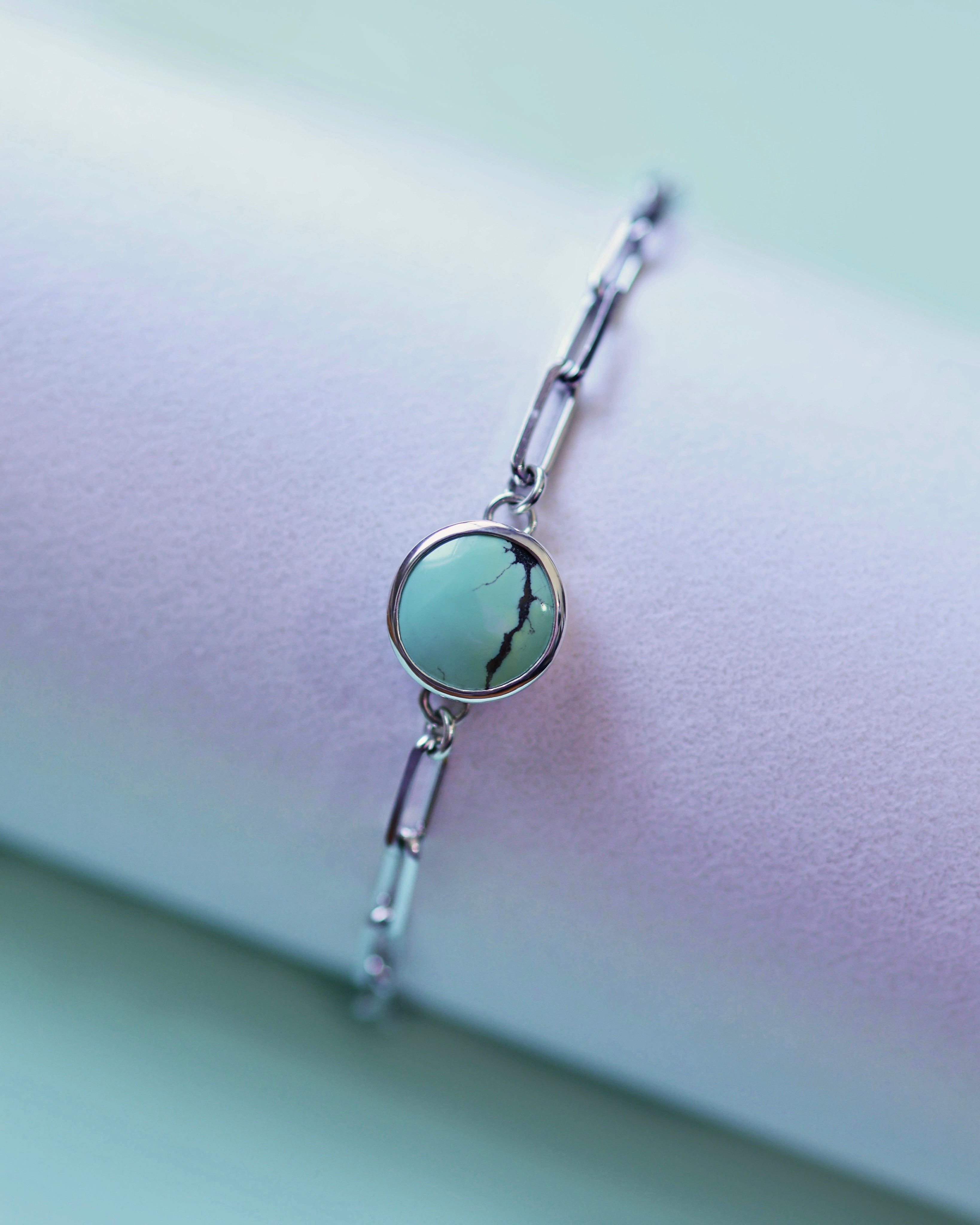 The Treasure Mountain Turquoise Bracelet No. 2 by Lilac Sky Studio is a sterling silver adjustable cable chain with a round turquoise stone featuring a dark vein, shown on a pastel-colored surface.