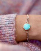 The Cloud Mountain Turquoise Bracelet No. 3 by Lilac Sky Studio, featuring a round turquoise stone on a silver chain, is shown on a wrist with a pink and purple textured sweater in the background.