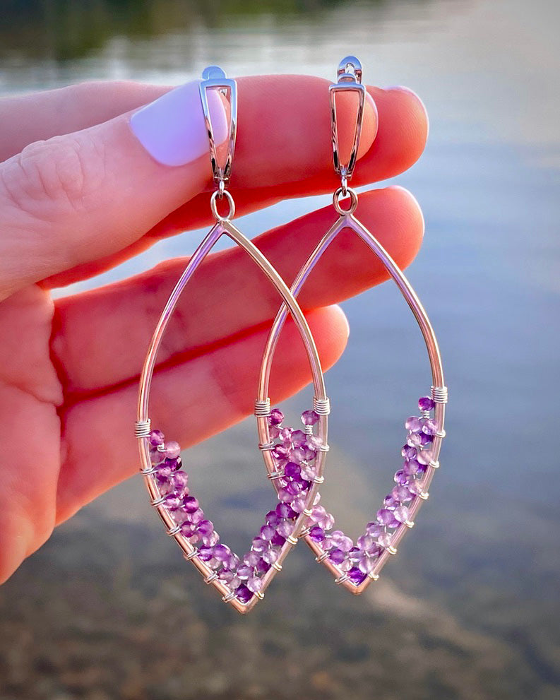 A hand displays Amethyst Earrings by Lilac Sky Studio—marquise-shaped sterling silver with clusters of handwoven amethyst beads—against a blurred lakeside background.